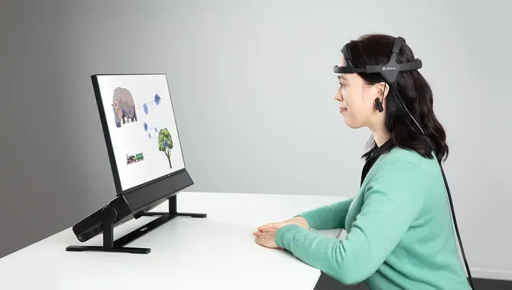 Woman wearing a virtual reality headset and headphones, sitting at a desk and watching a digital screen showing a cartoon image of a rhino, some fish, a tree, and a boat.