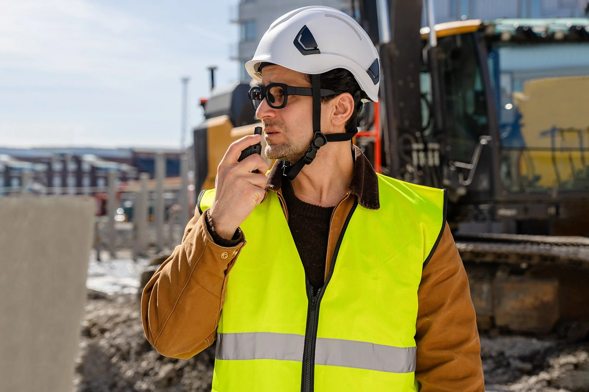 A male construction worker wearing a white safety helmet, black safety glasses, a brown jacket, and a bright yellow high-visibility vest, holding a walkie-talkie at a construction site with machinery and buildings in the background.