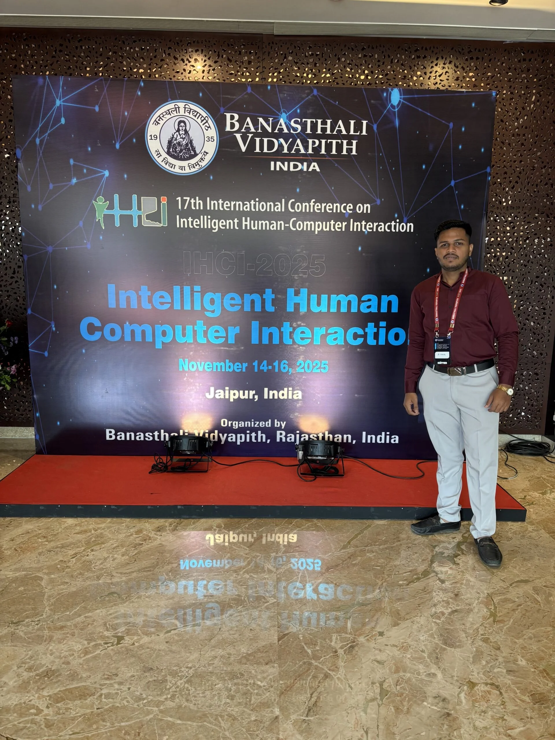 A man standing on a red carpet in front of a large digital display at a conference. The display reads: '17th International Conference on Intelligent Human-Computer Interaction, IHI-2025, November 14-16, 2025, Jaipur, India. The conference is organized by Banastali Vidyapith, Rajasthan, India.'