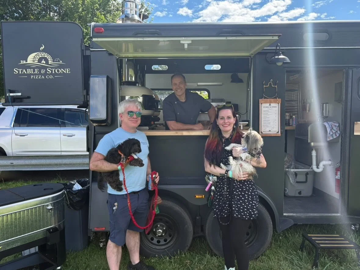 Three people with dogs in front of a dark-colored food truck labeled "Stable & Stone Pizza Co." on a sunny day in a grassy area.