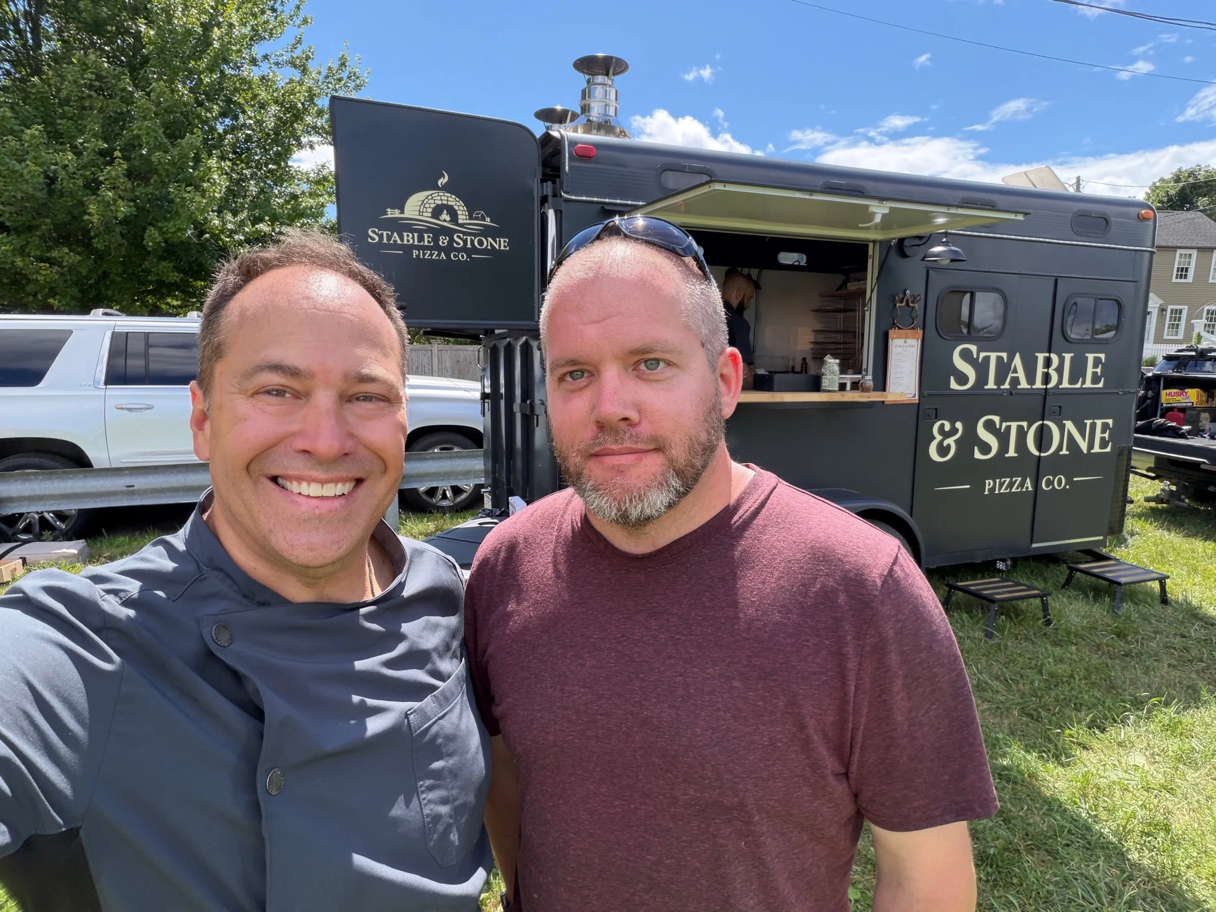 Two men taking a selfie in front of a food truck named 'Stable & Stone Pizza Co.' outdoors on a sunny day, with a tree and parked cars in the background.