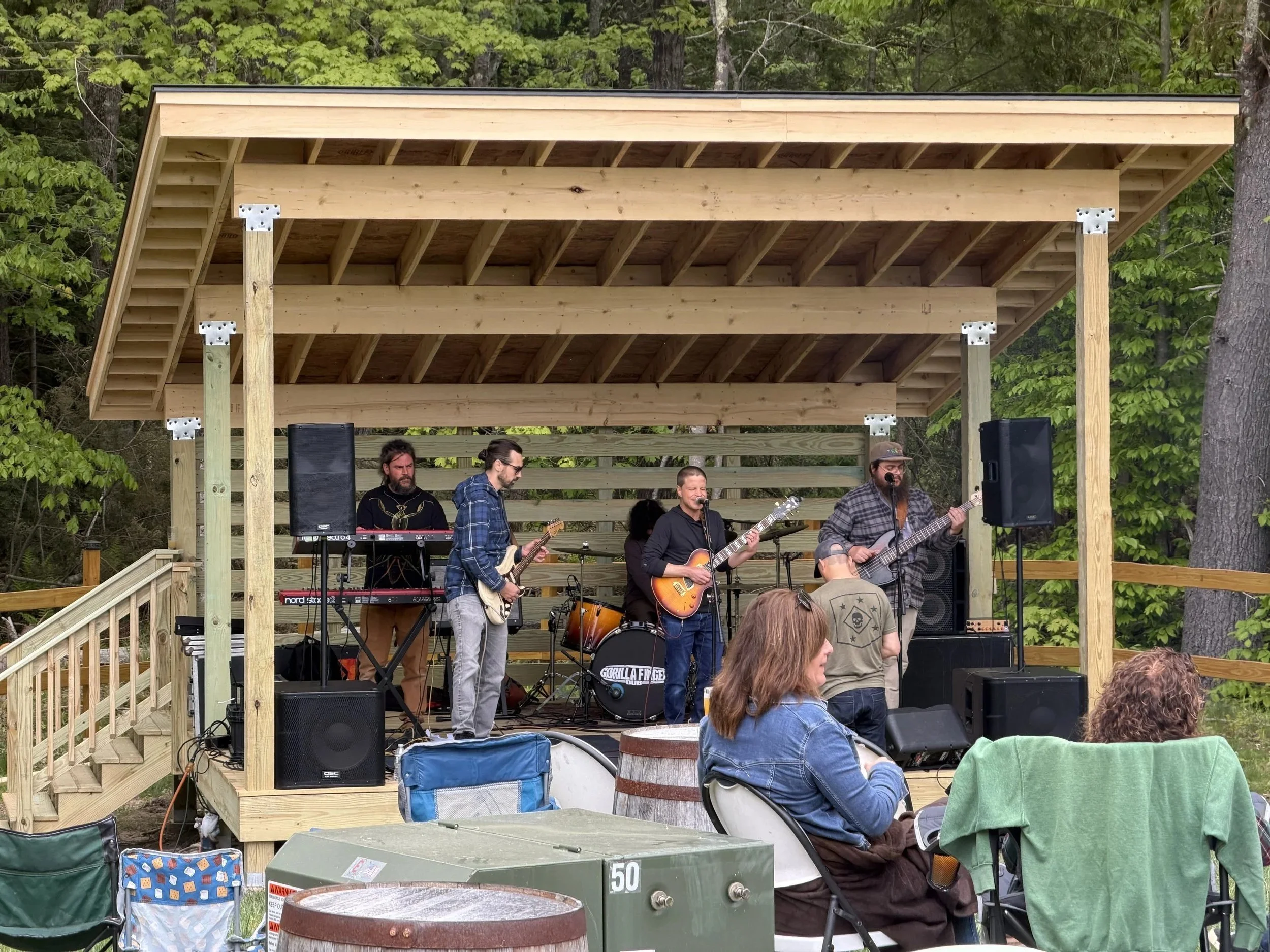 A band performs on an outdoor wooden stage in a forested area, with people seated in front, some wearing denim jackets and hats.