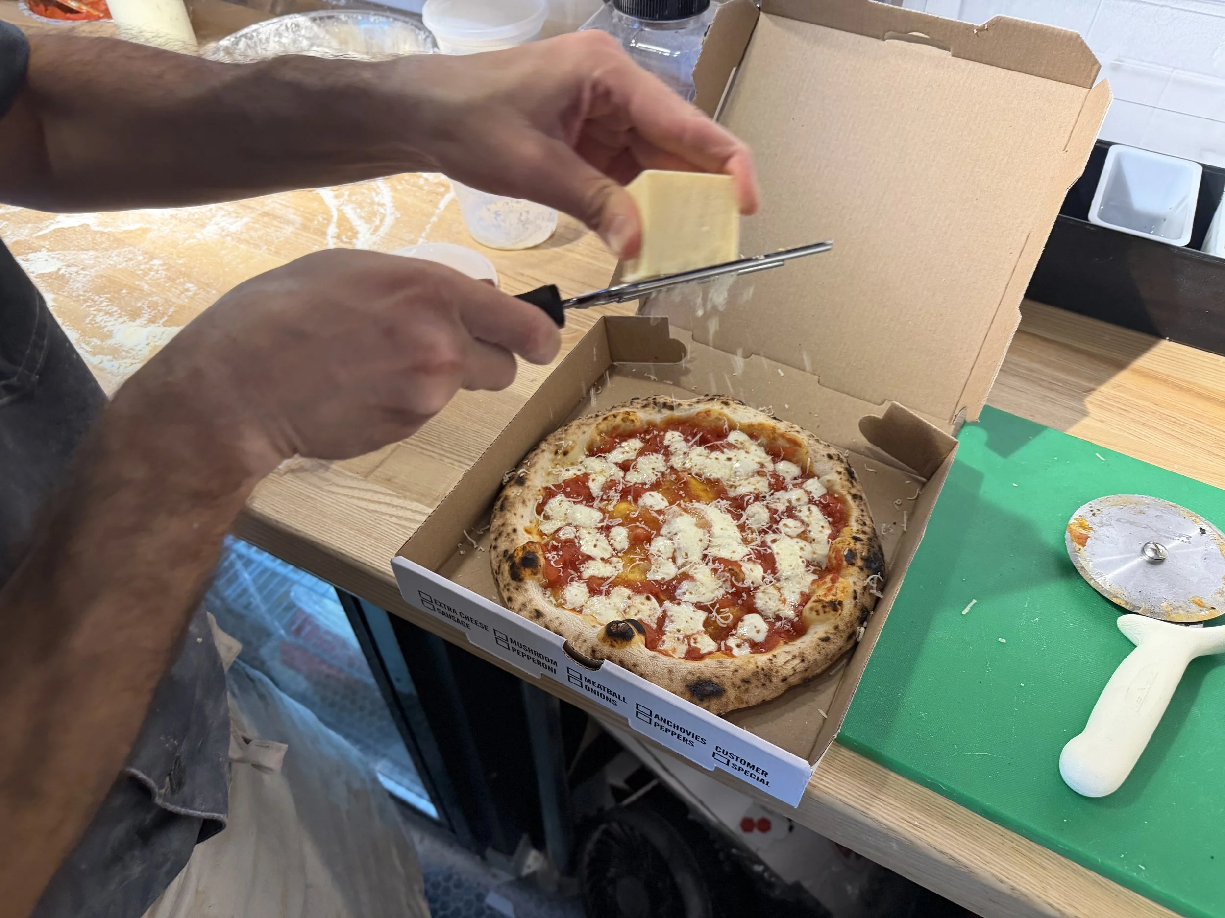 Person slicing a piece of cheese from a pizza in a takeout box on a table, with pizza toppings including cheese, tomato sauce, and possibly basil. There are various kitchen items and a green cutting mat on the table.