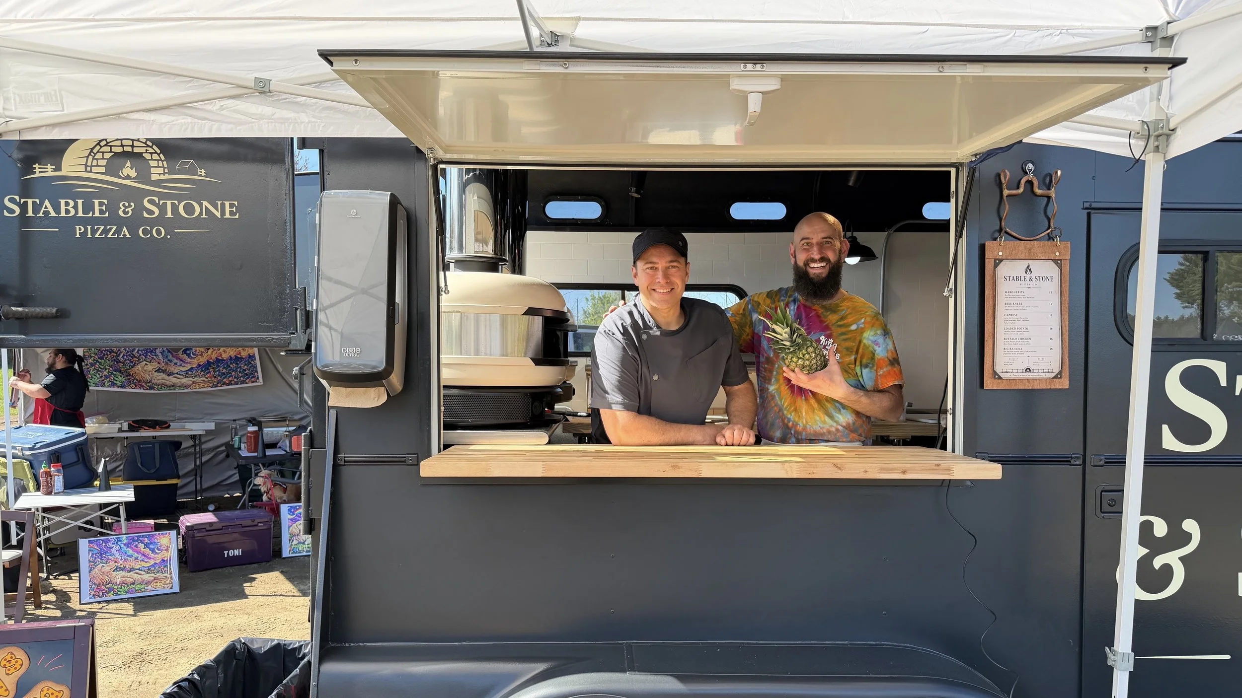 Two men stand inside a food truck labeled 'Stable & Stone Pizza Co.' smiling with one holding a pineapple. The truck has a menu on the right side and is set up outside at an event, with artwork and supplies visible in the background.