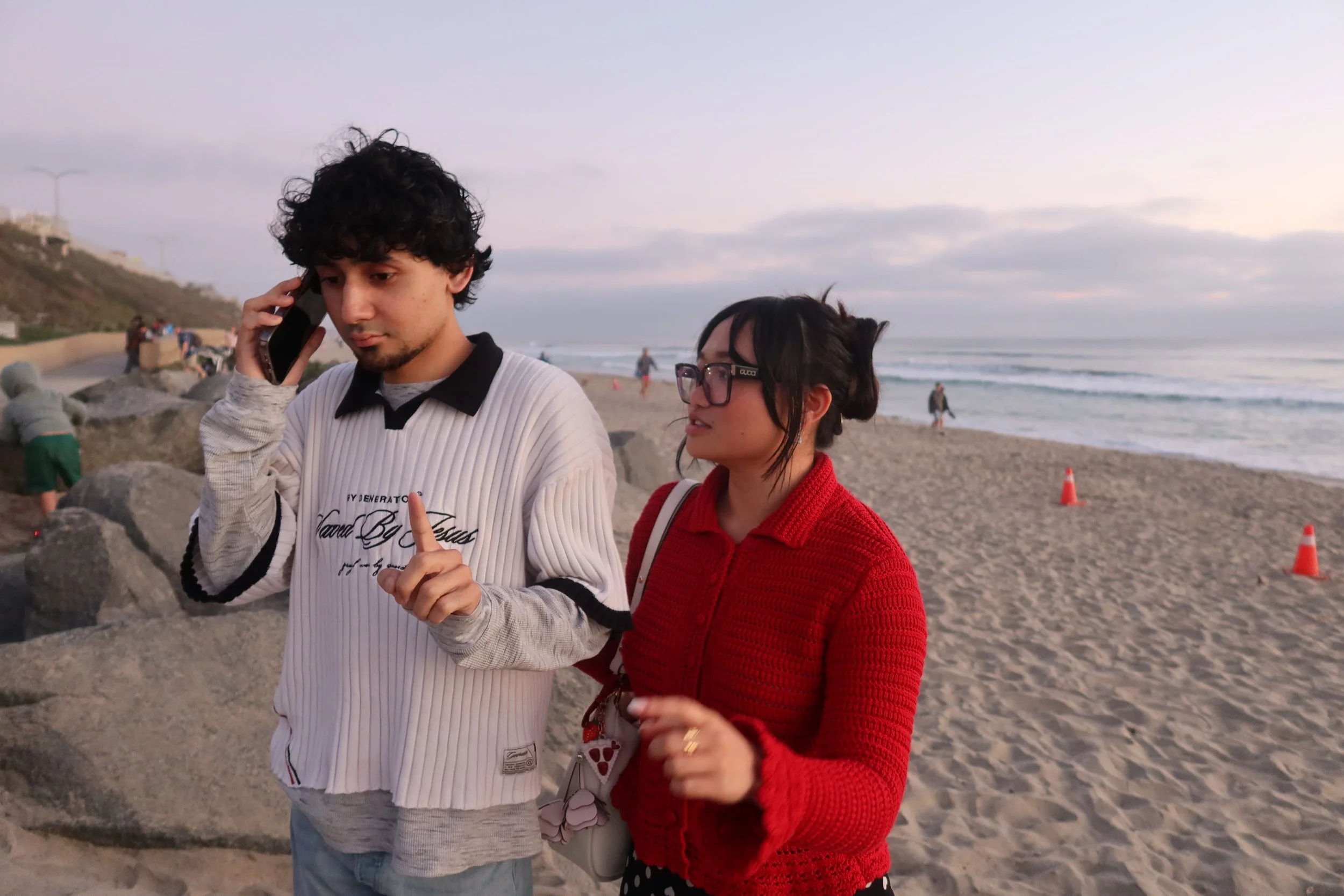 A young man with dark curly hair talking on a cell phone and a young woman with glasses and dark hair wearing a red sweater standing on a beach with waves and other people in the background.