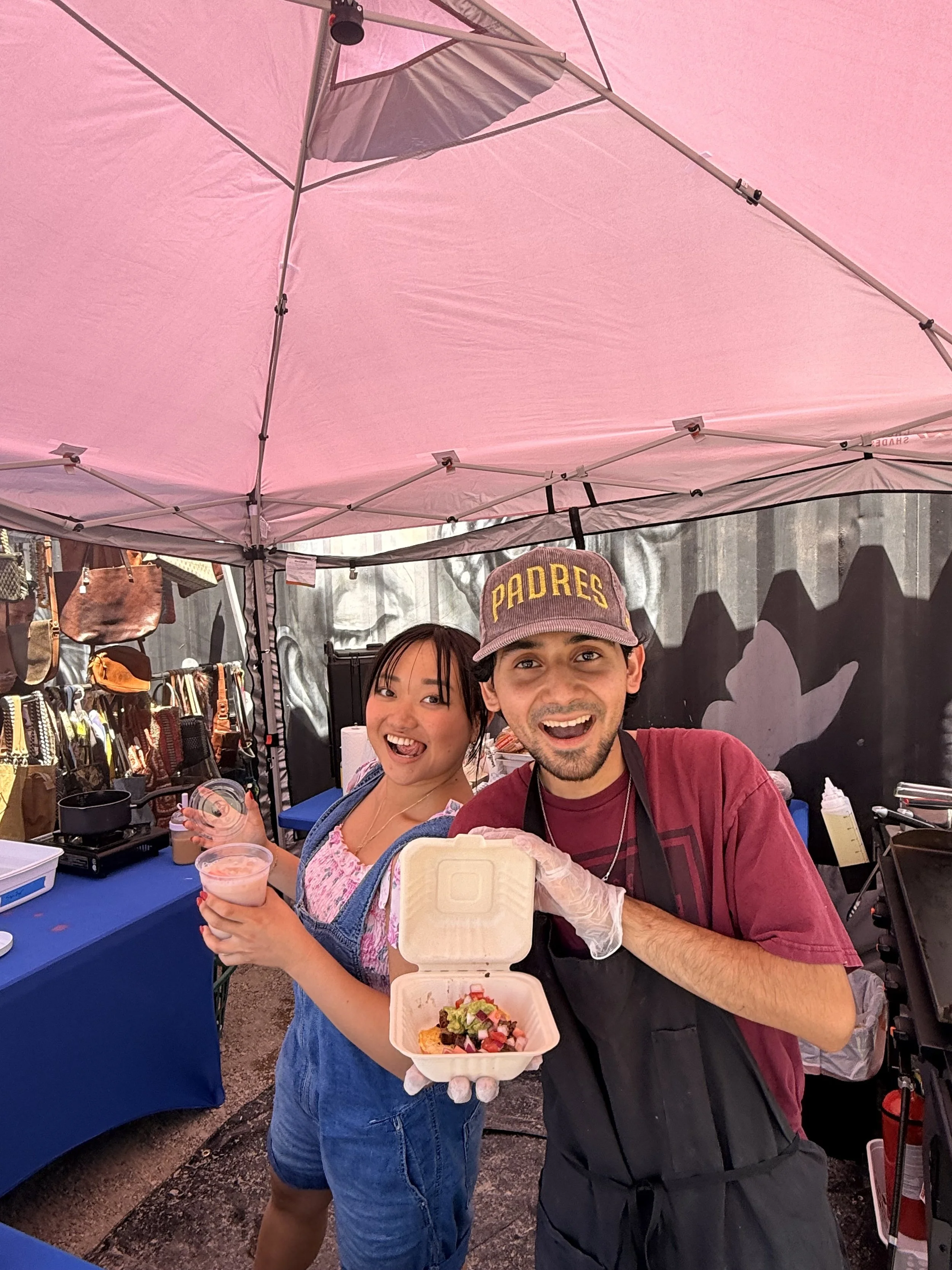 Two people are smiling and posing at a food stall at an outdoor event, holding a food container with a salad. They are under a pink canopy, with various bags and merchandise displayed on the side. The woman on the left is wearing a pink top, and the man on the right is wearing a cap with the word 'Padres' on it and a maroon shirt, with a black apron and gloves.