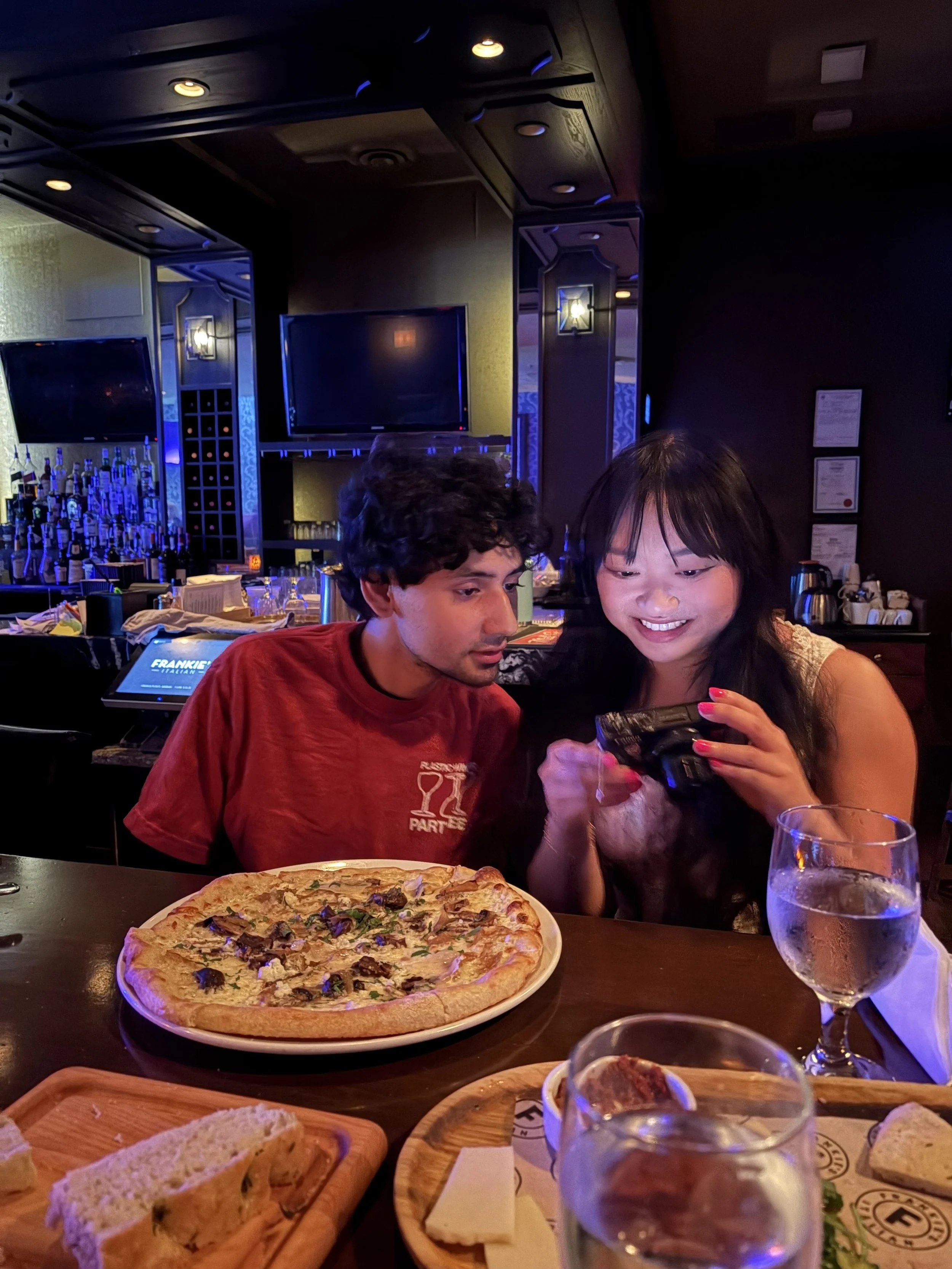A pizza with toppings on a plate, a glass of water, a wooden board with bread, and two people smiling at a dinner table in a dimly lit restaurant.