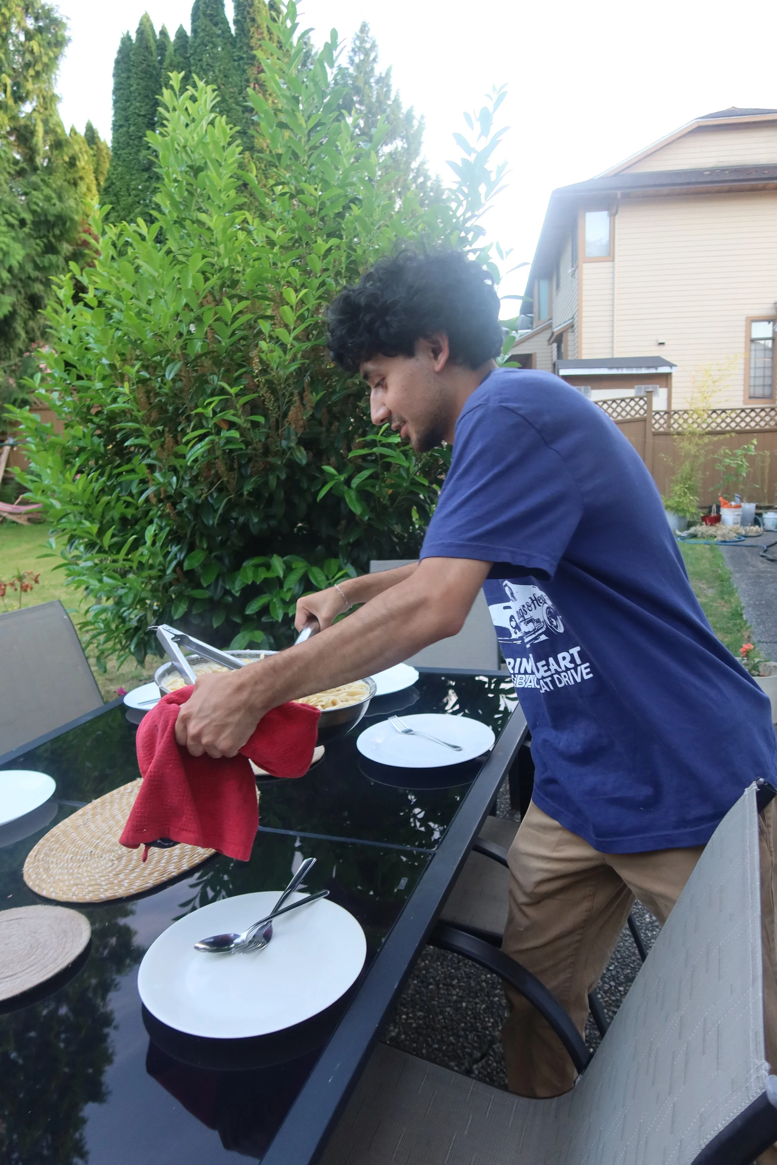 A man in a blue T-shirt and tan shorts is standing outside at a dining table, serving food into a bowl. The table is set with white plates, silverware, and woven placemats, surrounded by chairs. There is a lush green garden with bushes and trees in the background, with a house visible behind the garden.