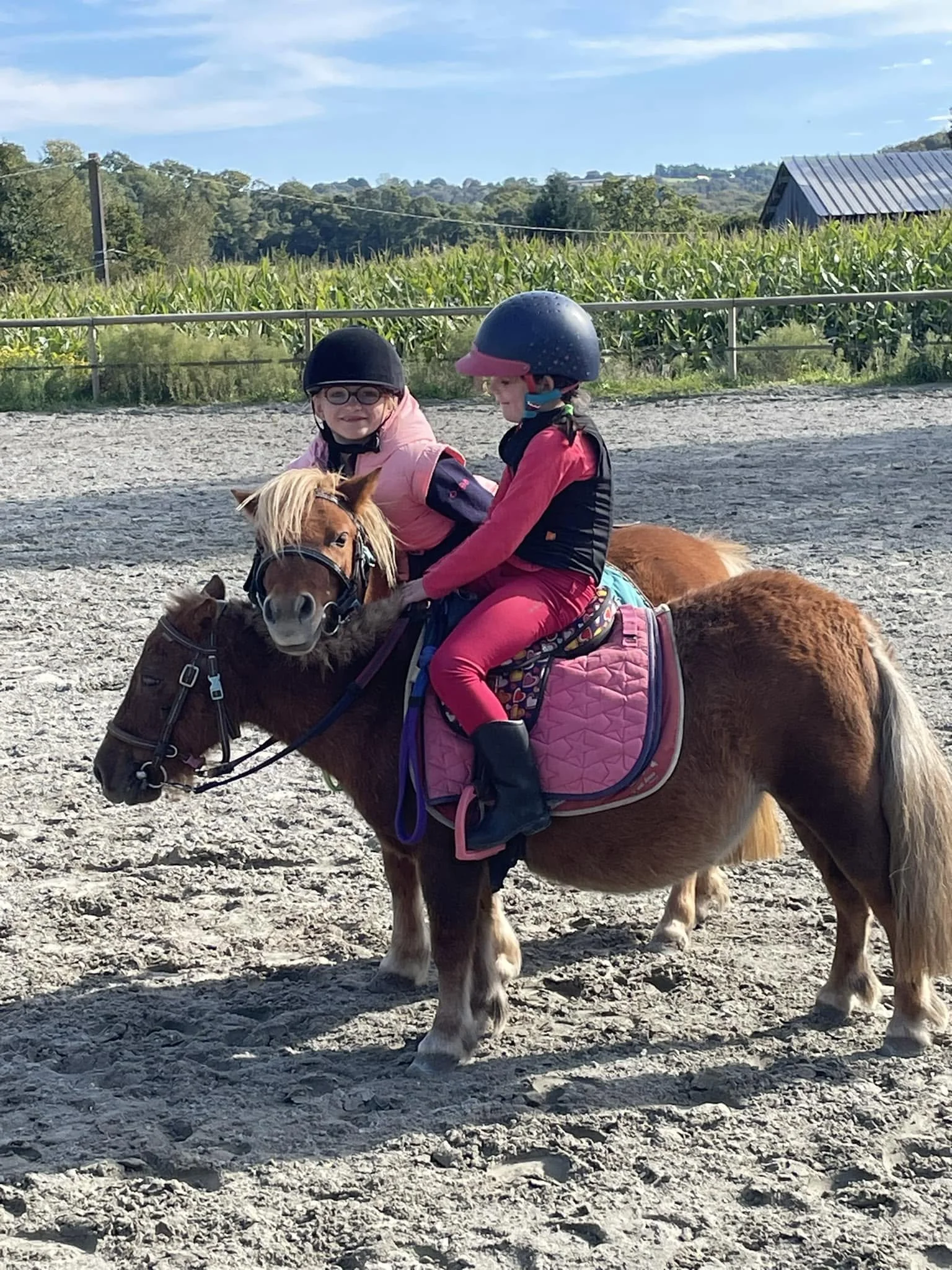 Deux jeunes filles montent un poney dans un enclos extérieur, portant des casques de protection et de vêtements colorés.