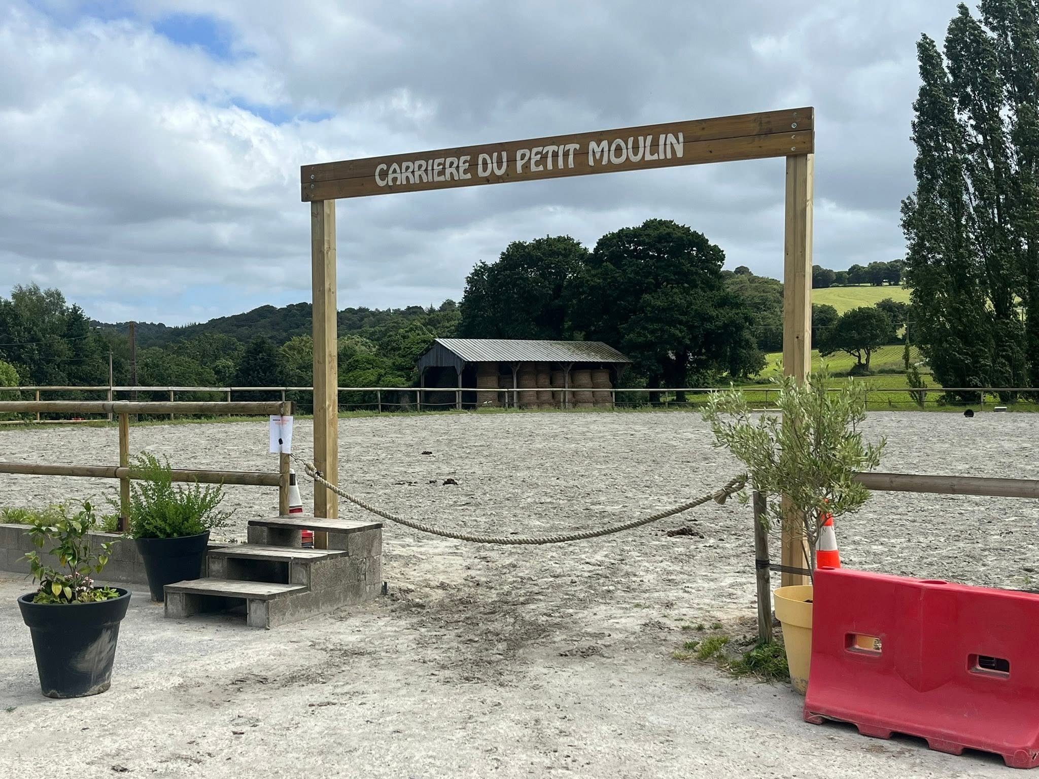 Un panneau en bois indique 'Carrière du Petit Moulin'. L'arrière-plan dévoile un paysage rural avec des collines et des arbres. Au premier plan, il y a des pots de fleurs, une petite structure en bois, une barrière, et quelques éléments de signalisation.