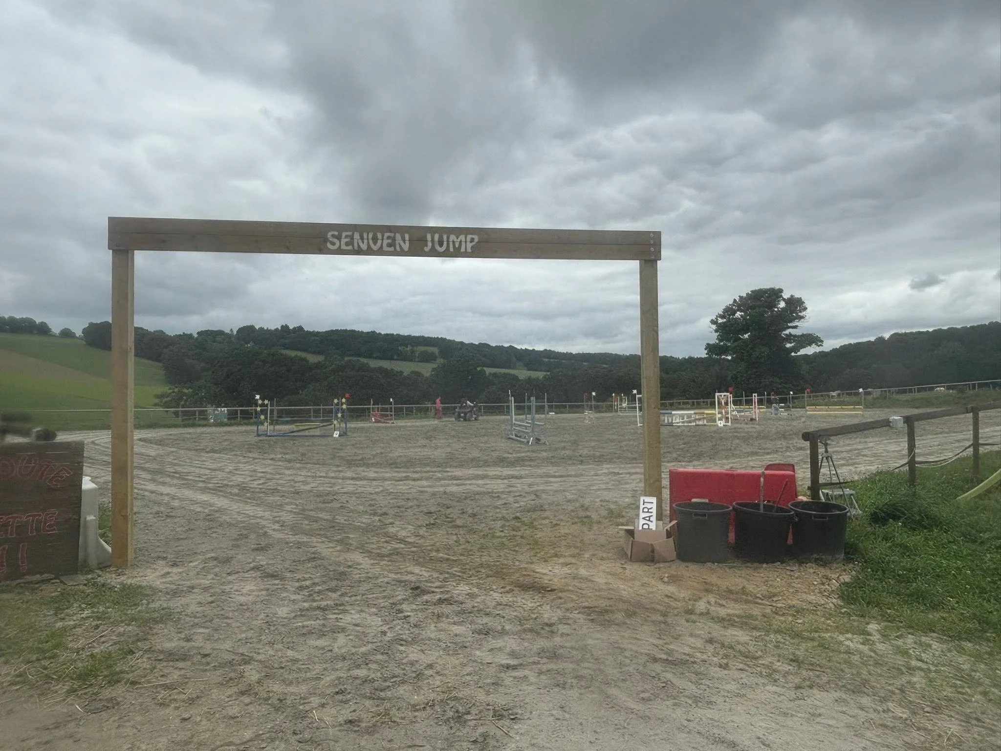 Une vue d'un espace d'équitation extérieur avec des obstacles pour le saut d'obstacles, un panneau indiquant le départ, et un ciel nuageux sur des collines verdoyantes en arrière-plan.