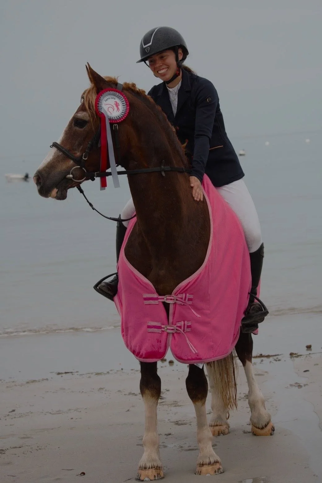 Une jeune femme en casque et en tenue d'équitation sourit, montant un cheval sur la plage, portant une couverture rose autour du corps de l'animal, avec un ruban de victoire attaché à la tête du cheval.