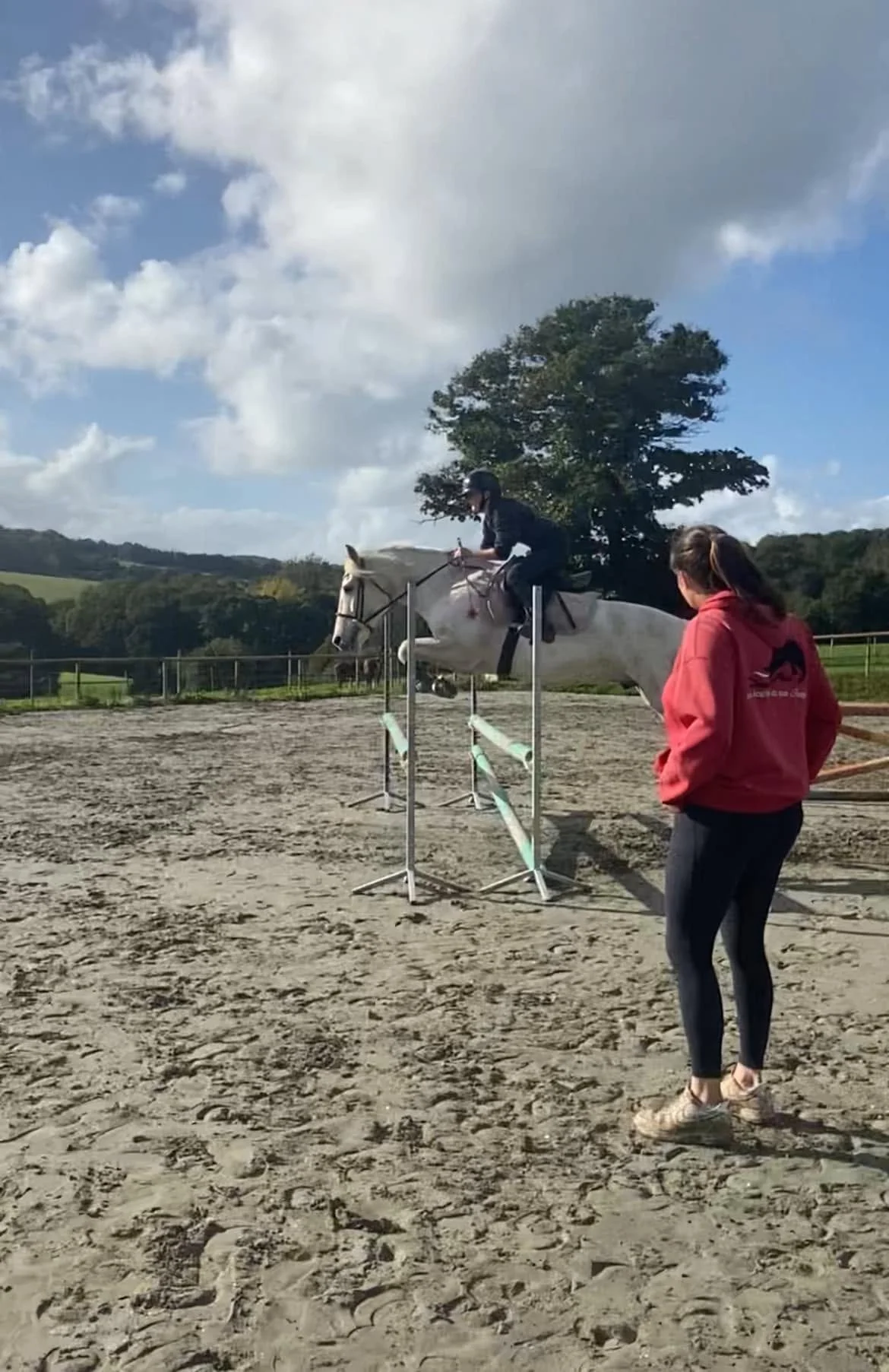 Une personne fait passer un cheval blanc lors d'un saut d'obstacles dans un centre équestre, avec une autre personne observant, sur un fond de paysage rural et un ciel partiellement nuageux.