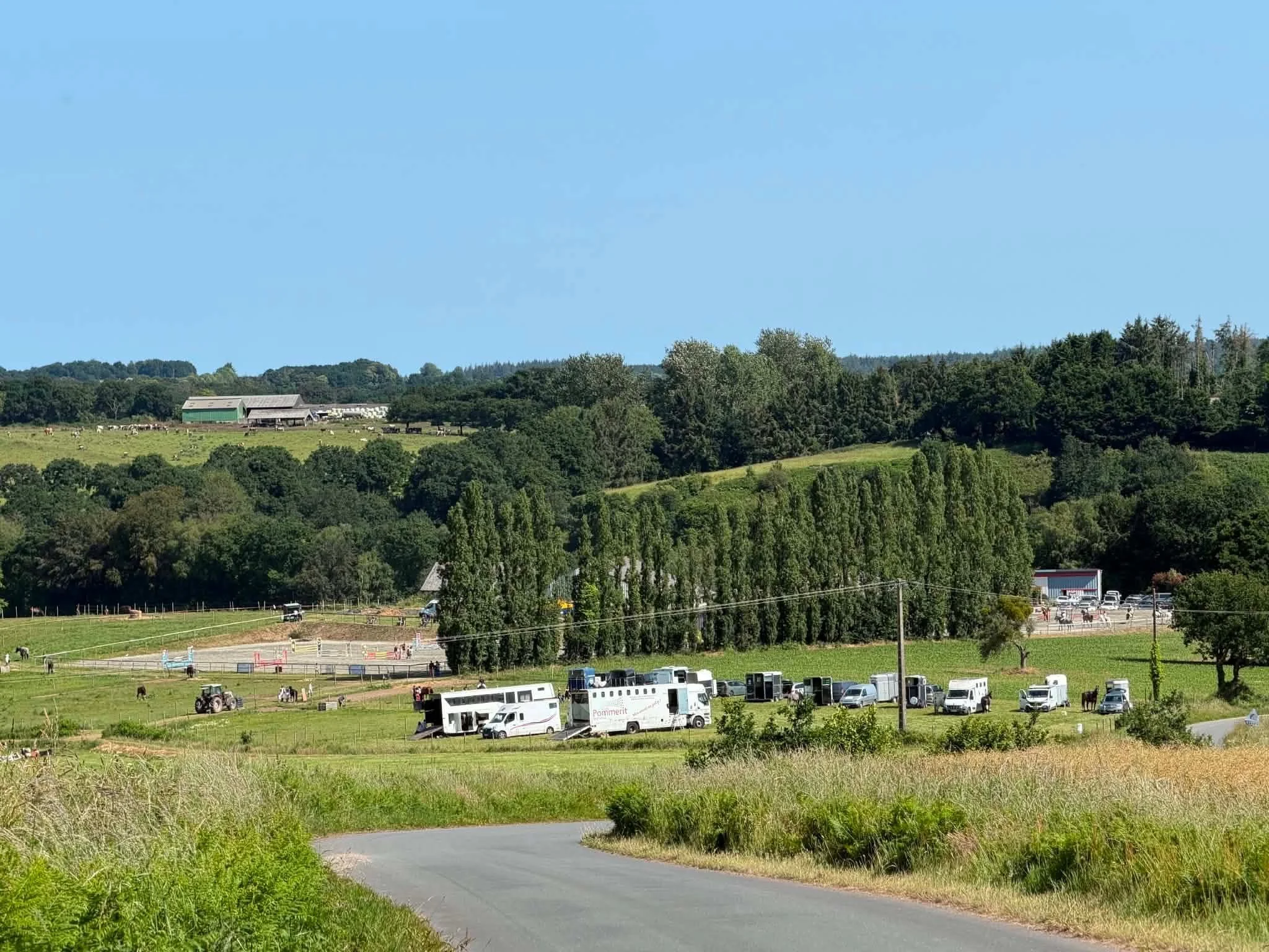 Paysage rural avec prairies, arbres, ferme et véhicules, ciel bleu.