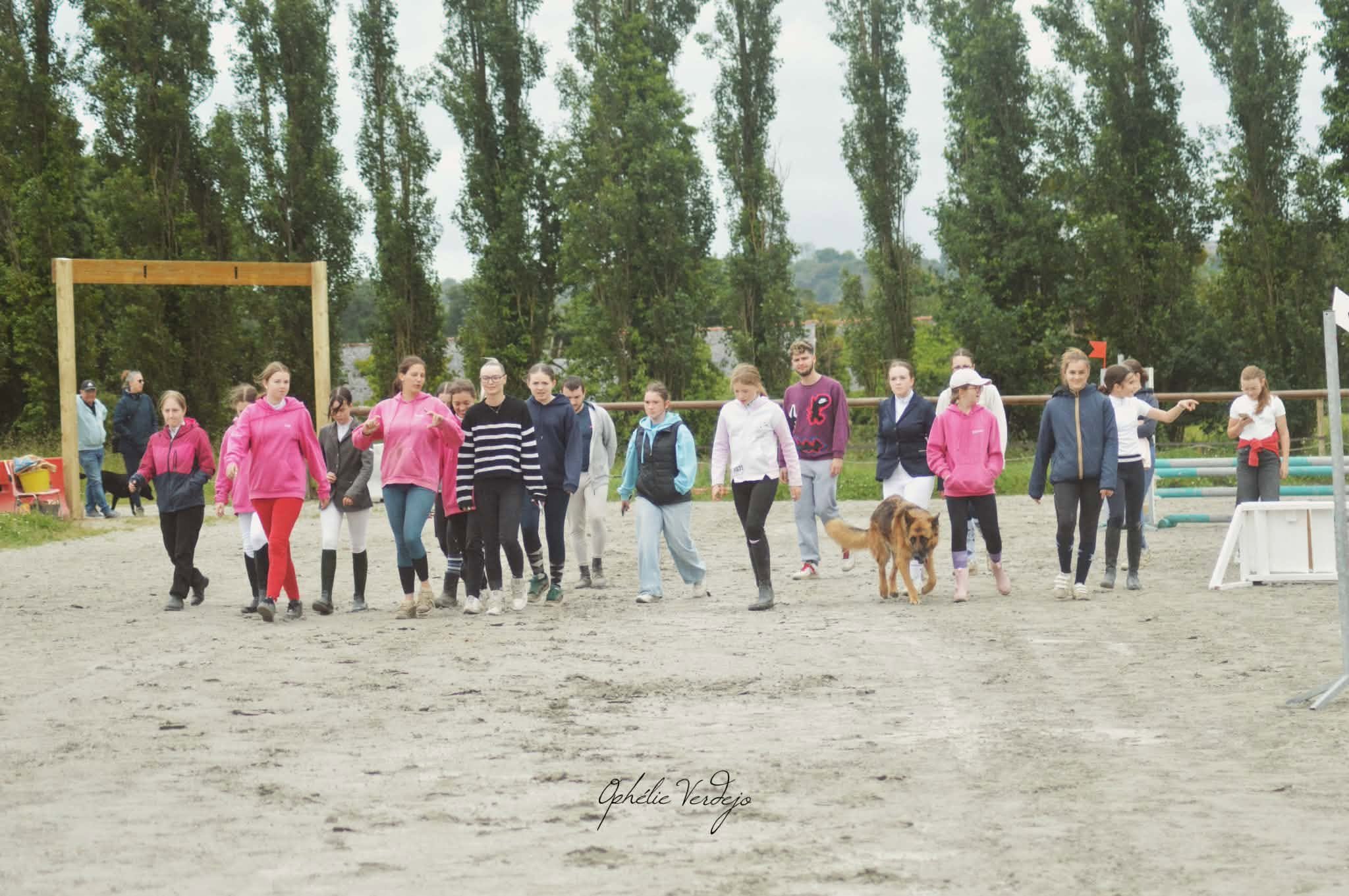 Un groupe de personnes, principalement des jeunes filles, marche ensemble dans un terrain d'équitation, accompagnées de chiens, sous un ciel nuageux, avec des arbres en arrière-plan.