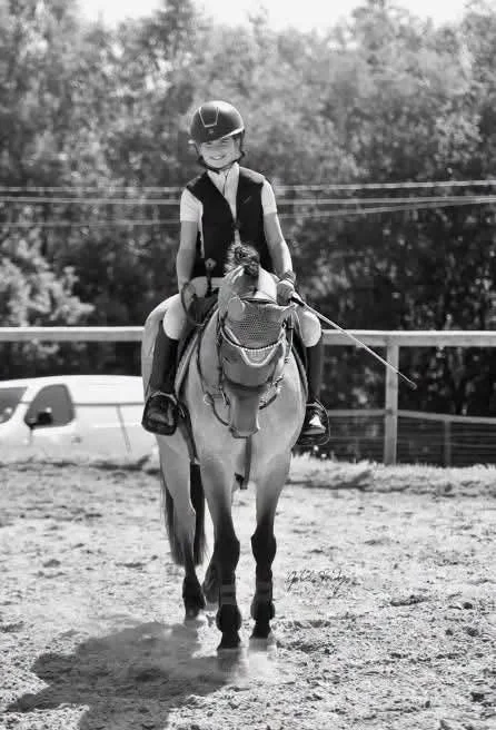 Une jeune fille en équipement d'équitation, portant un casque, assise sur un cheval dans un espace extérieur avec des arbres en arrière-plan.