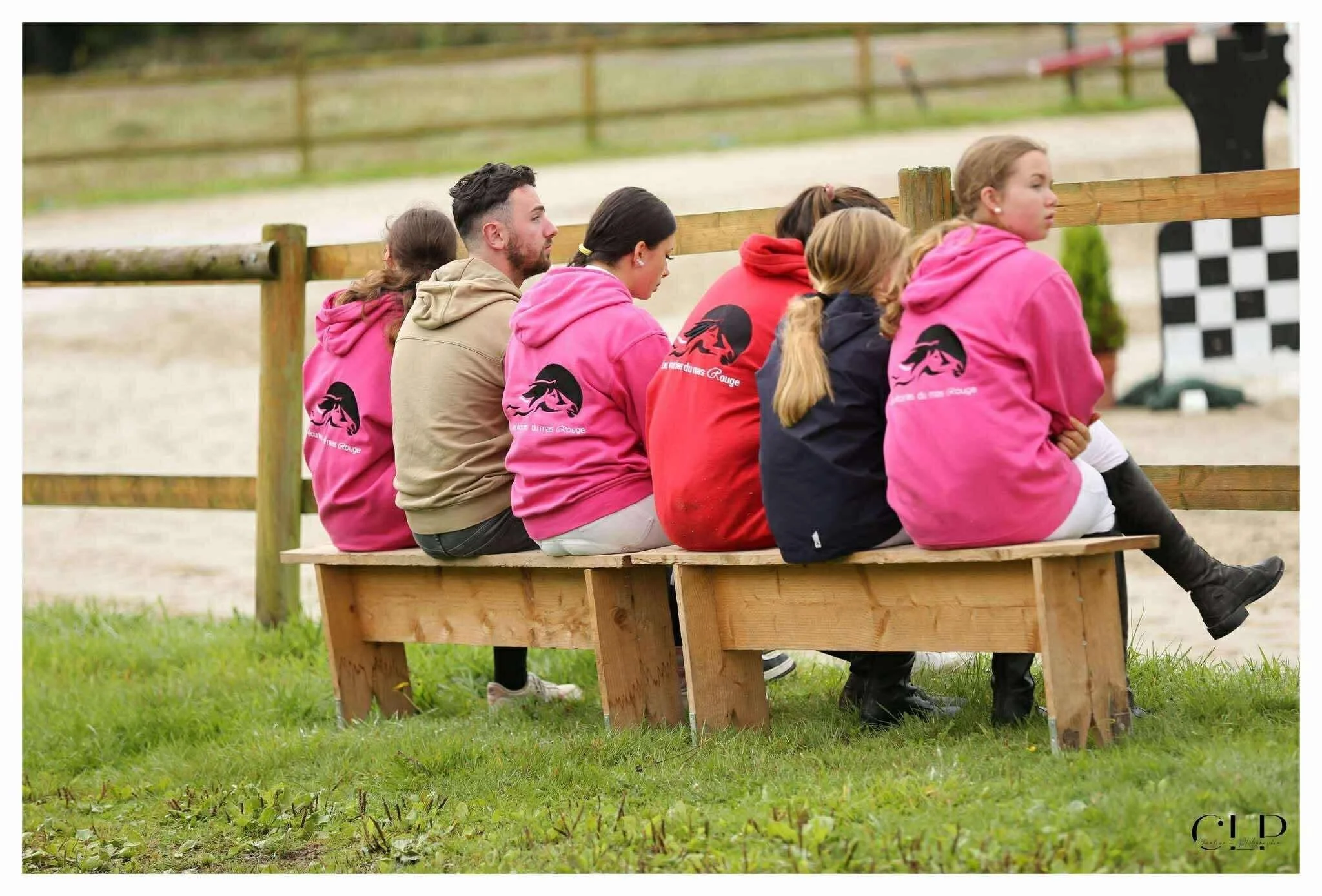 Groupe de jeunes assis sur un banc en bois à l'extérieur, portant des pulls roses, un sous un beige, dans un lieu de sport ou de loisir. Fond naturel avec un pré et une clôture en bois.