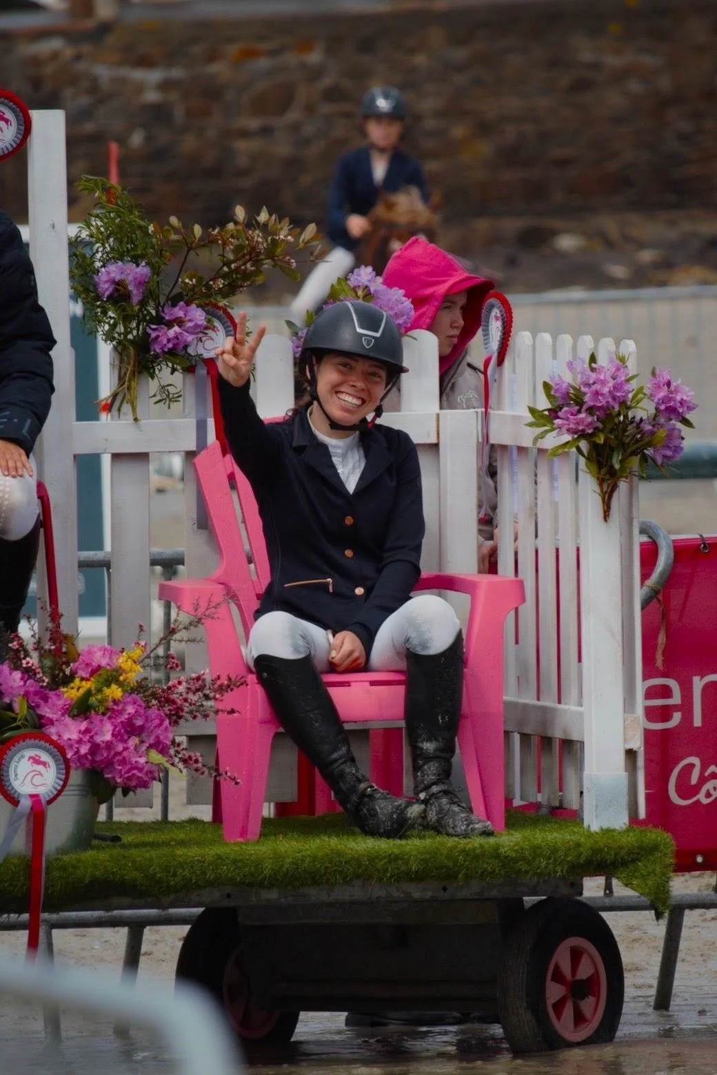 Une jeune cavalière souriante, portant un casque noir, assise sur une chaise rose en haut d'une charrette décorée de fleurs, avec une autre personne à l'arrière portant une capuche rose. La scène semble avoir lieu lors d'une compétition équestre.