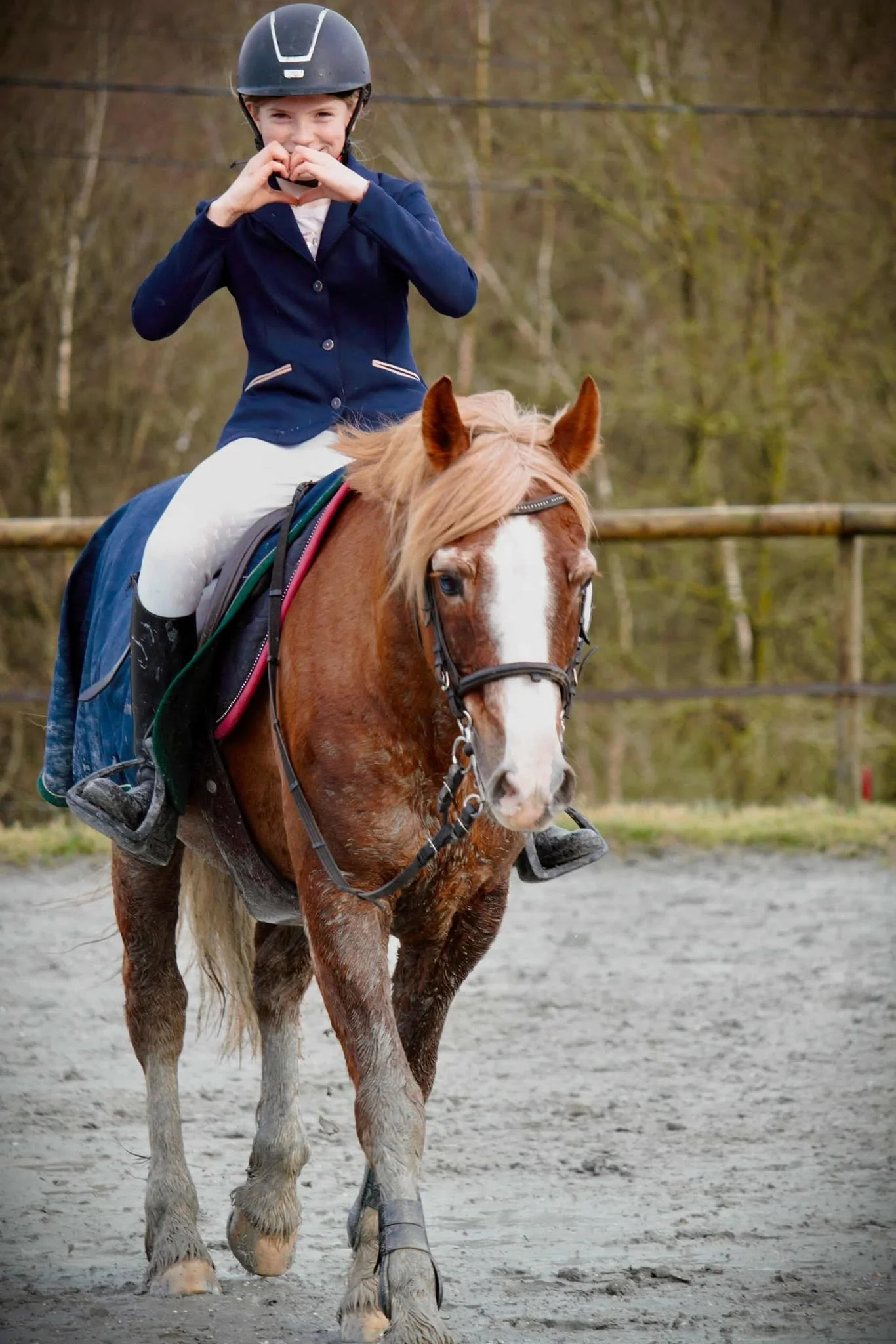 Une jeune fille souriante porte un casque à cheval et fait un signe de cœur avec ses mains. Elle est assise sur un cheval dans un enclos en extérieur, avec des arbres en arrière-plan.