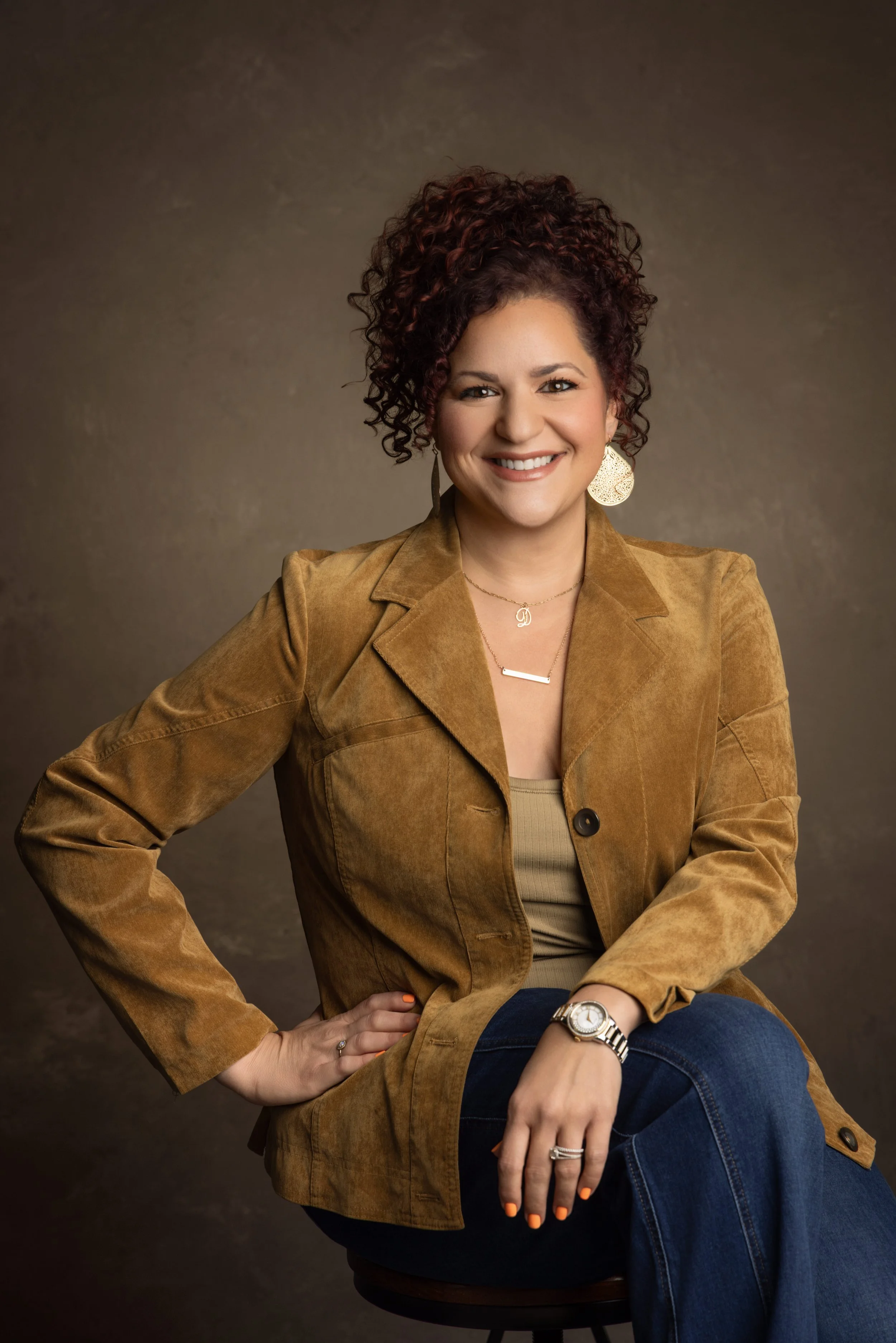 A woman with curly red hair sitting on a stool, smiling, wearing a brown jacket, gold jewelry, and blue jeans against a dark background.