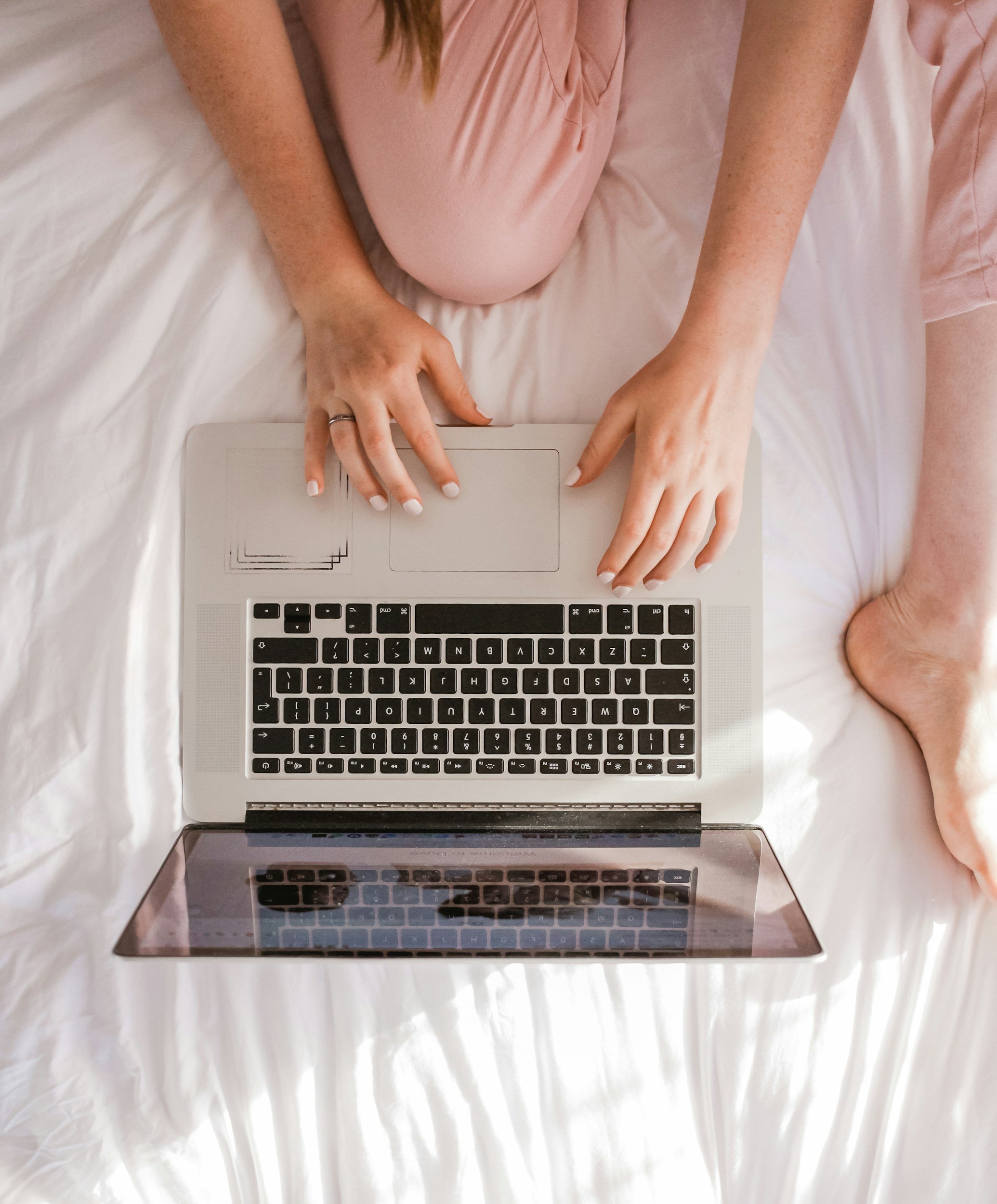 A person with freckled arms, wearing pink pajamas, sitting on a white bed, using a silver laptop.
