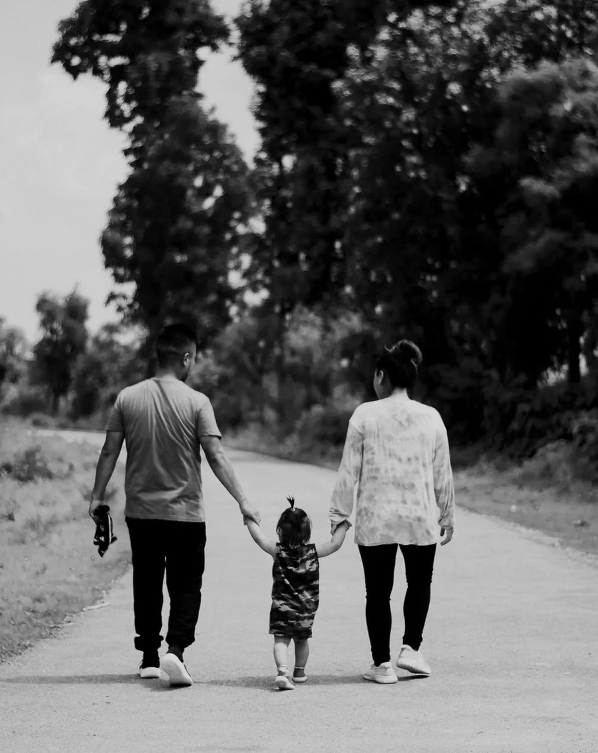 A black-and-white photo of a family walking hand-in-hand on a rural road. The family consists of two adults and a young child, with trees lining the road in the background.