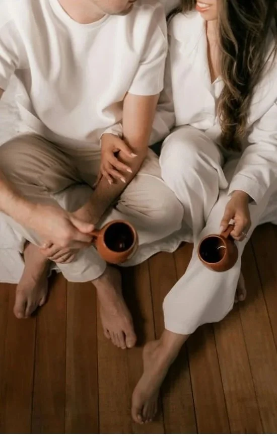 Close-up of a couple sitting on a wooden floor, holding mugs of coffee, with their legs and hands intertwined, smiling at each other.
