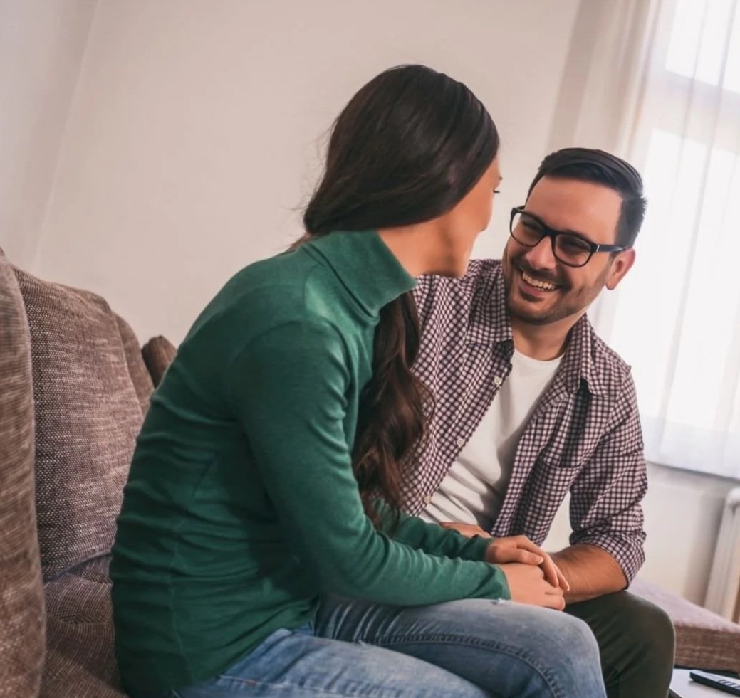 A man and woman sitting on a couch, holding hands, smiling and engaging in conversation in a cozy, well-lit room.