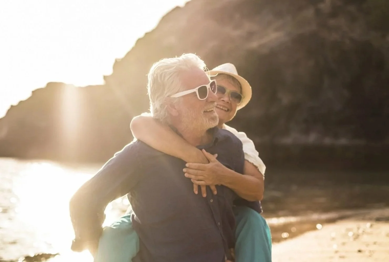 An elderly man and woman enjoying a beach sunset, with the woman giving a piggyback ride on the man's back. Both are smiling, wearing sunglasses, and the woman has her arms around the man's neck. The scene is backlit by the setting sun, with rocky cliffs and water in the background.
