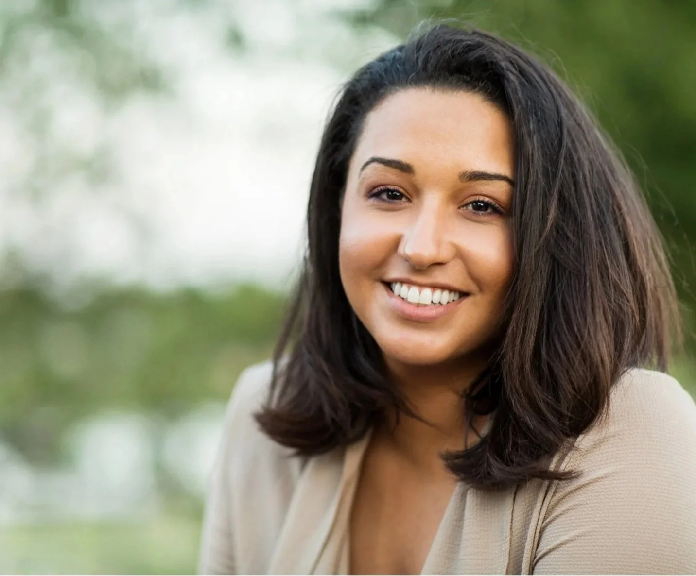 A smiling woman with shoulder-length dark hair, wearing a light-colored top, outdoors with blurred greenery in the background, trusted virtual therapist in New Jersey