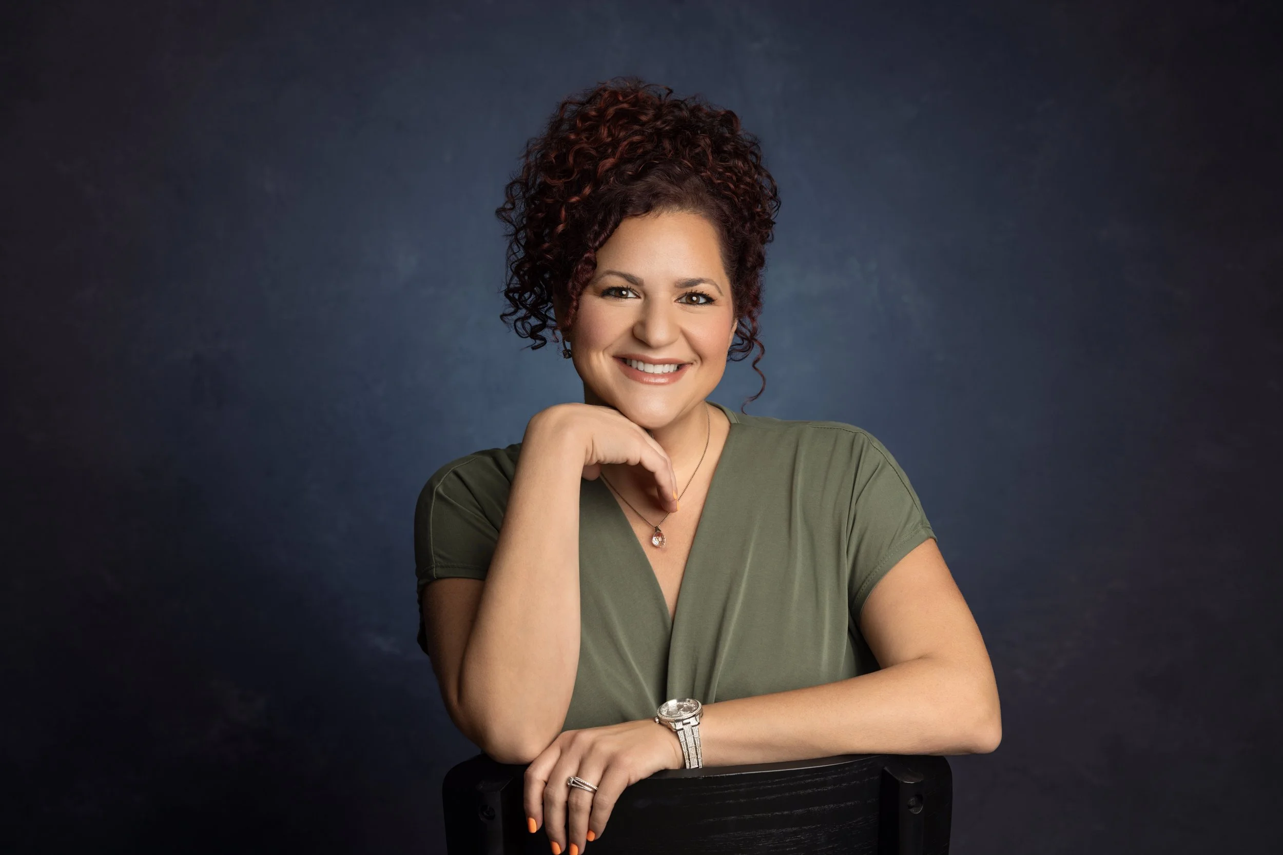 A woman with curly, reddish-brown hair styled up, smiling with her hand resting on her chin, wearing a green top, watch, and necklace, sitting against a dark background.