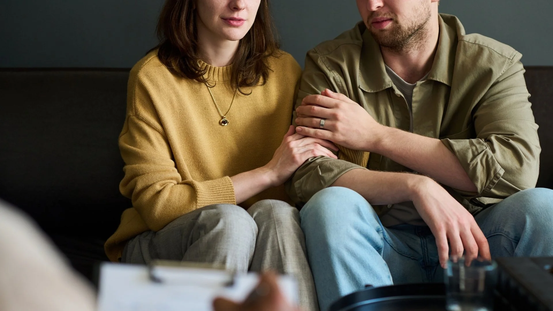 A man and woman sitting closely on a couch, holding hands, with the woman's hand on the man's arm. Both are casually dressed, and the scene conveys intimacy and support.