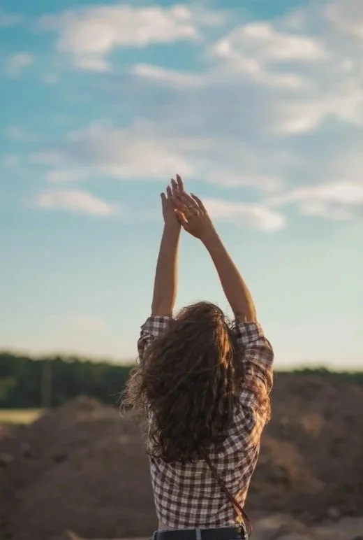 A woman with curly hair wearing a checkered shirt standing outdoors with arms raised toward the sky, during sunset or late afternoon, with a sky and landscape background.