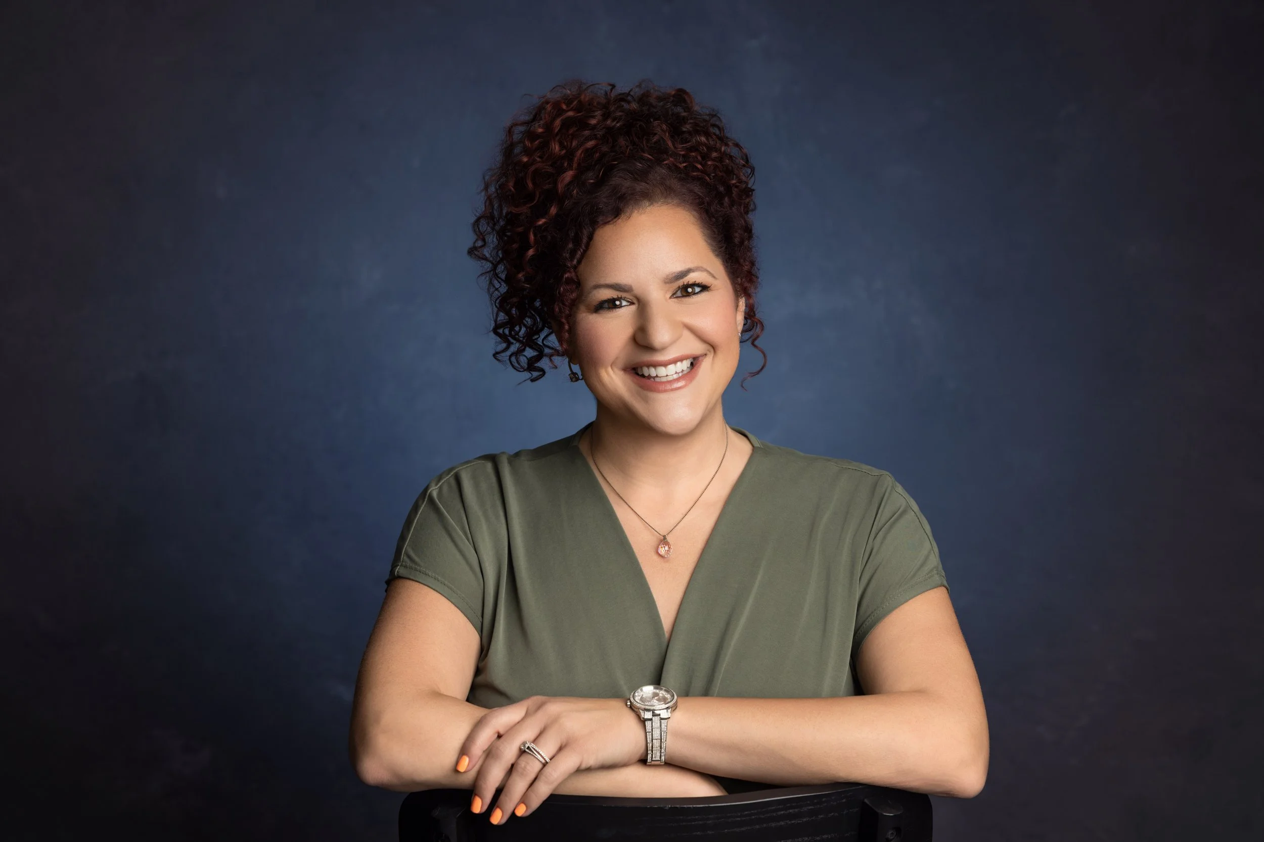 A woman with curly hair and a bright smile, wearing a green shirt, a watch, and jewelry, crossing her arms, against a dark background.