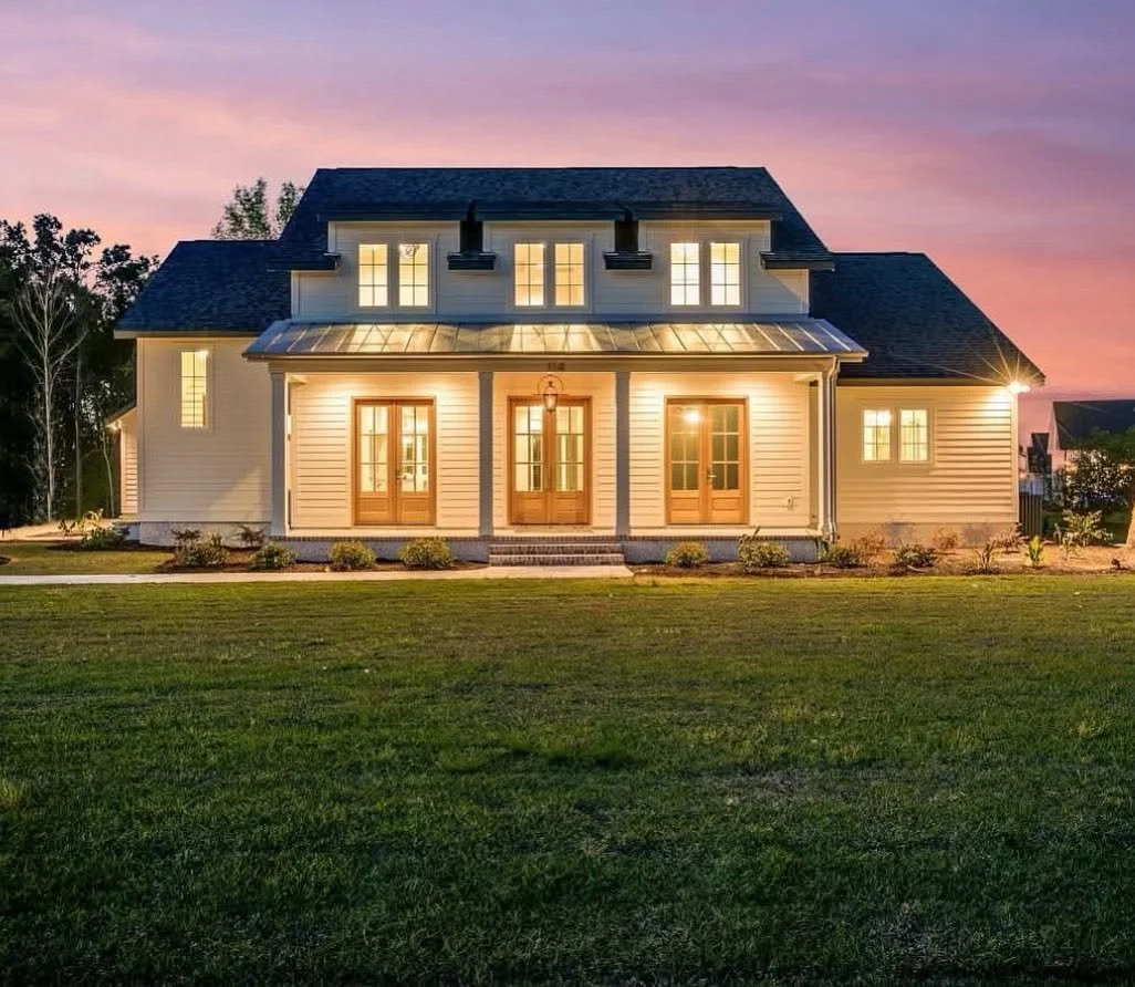 A large, two-story house with white siding and multiple windows, illuminated at dusk with a purple and pink sky.