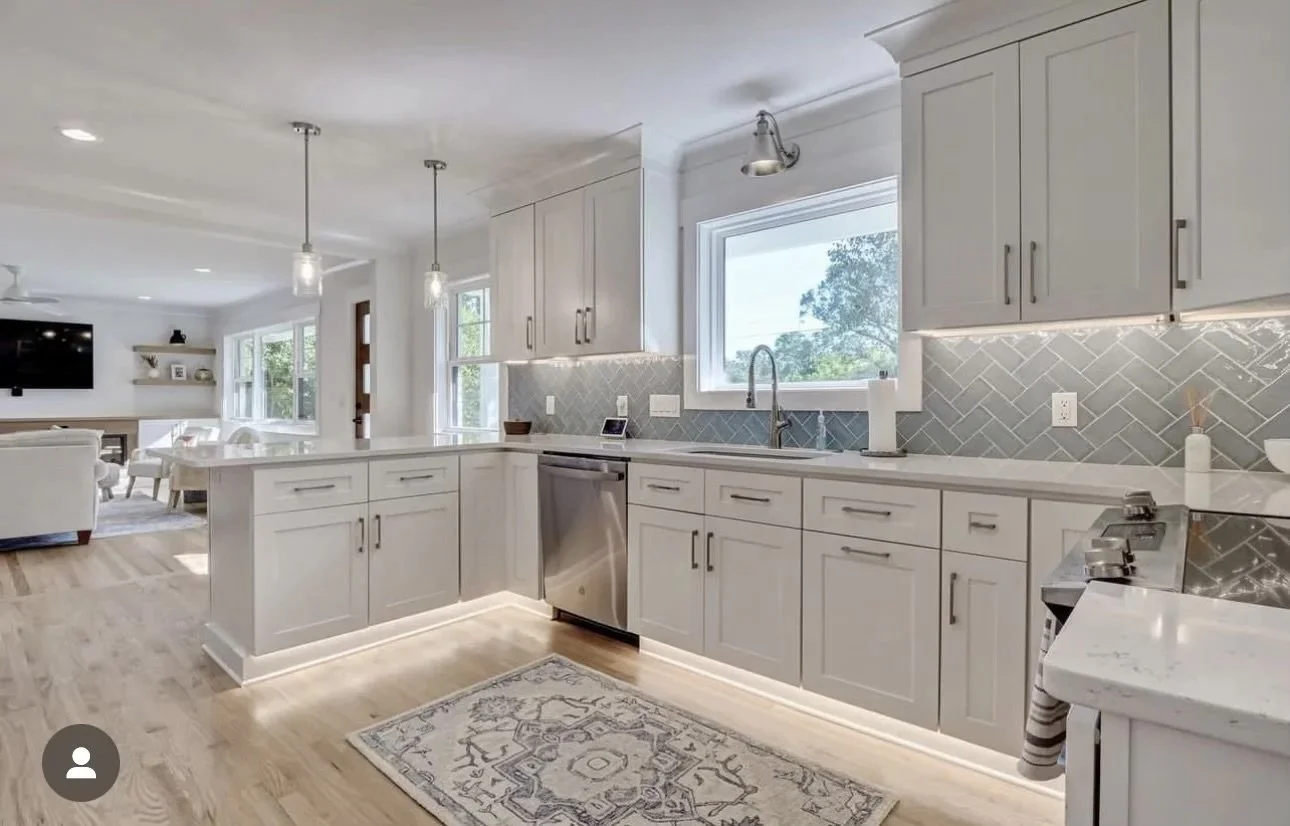 Modern kitchen with white cabinets, gray tile backsplash, and stainless steel appliances. There is a large window above the sink, and a living room is visible in the background.