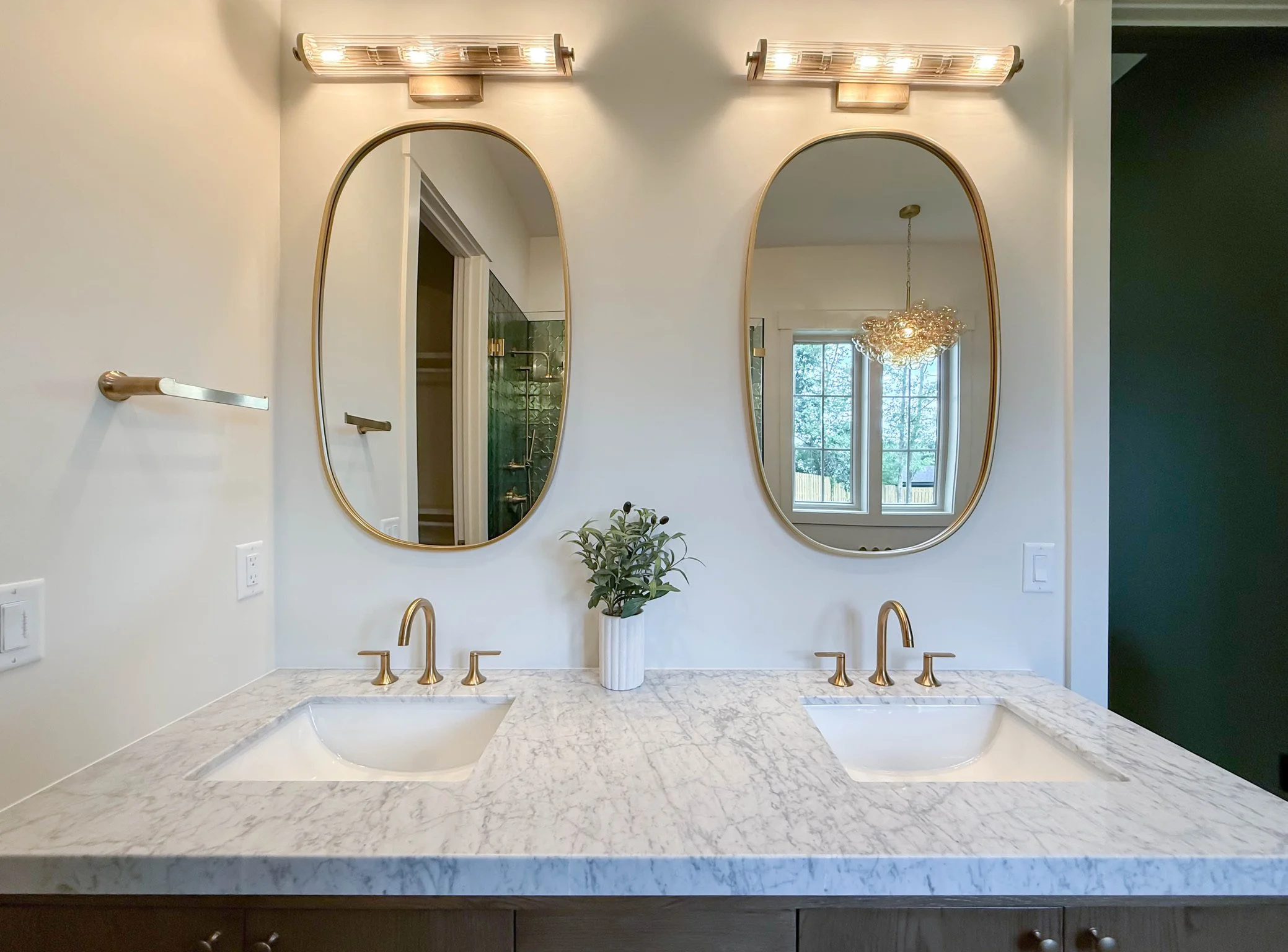 A bathroom vanity with a marble countertop, double sinks with gold faucets, and a pair of oval mirrors framed in gold. There are wall-mounted gold light fixtures above each mirror and a potted plant in between. A window and dining chandelier are reflected in the mirrors.