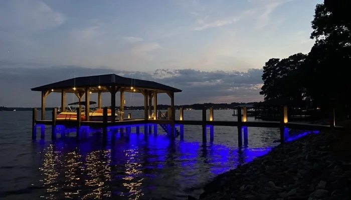 A lakeside dock with a covered seating area and blue lighting reflections on the water at dusk, with trees in the background.