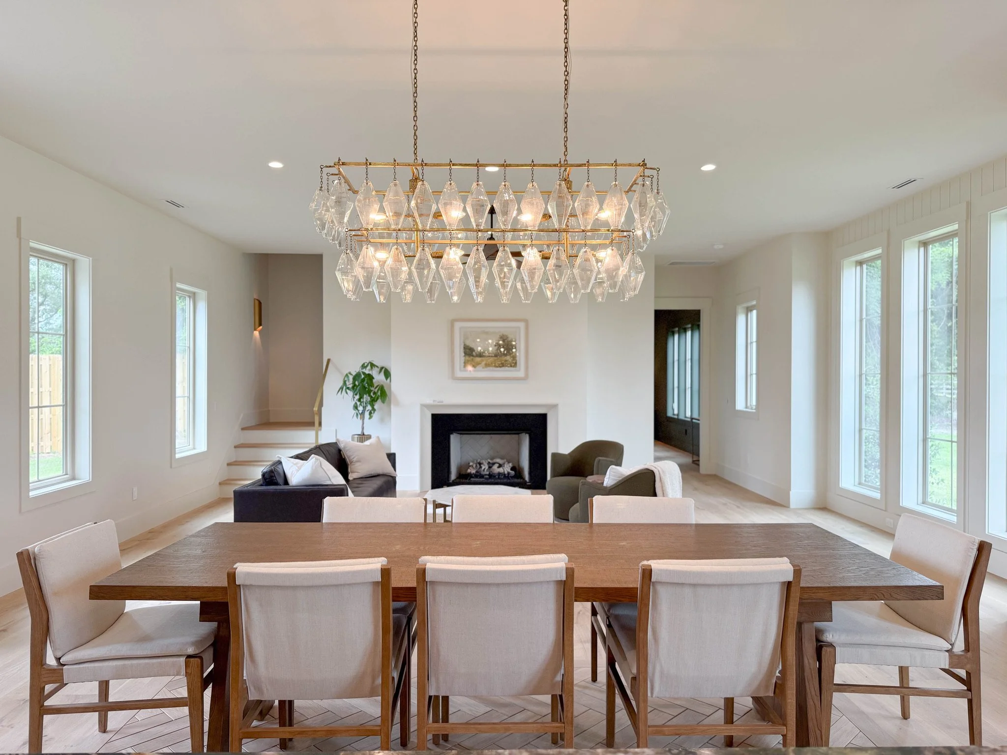 Dining room with a large wooden table surrounded by beige upholstered chairs, a modern chandelier with glass and gold accents hanging above, and multiple windows allowing natural light in.