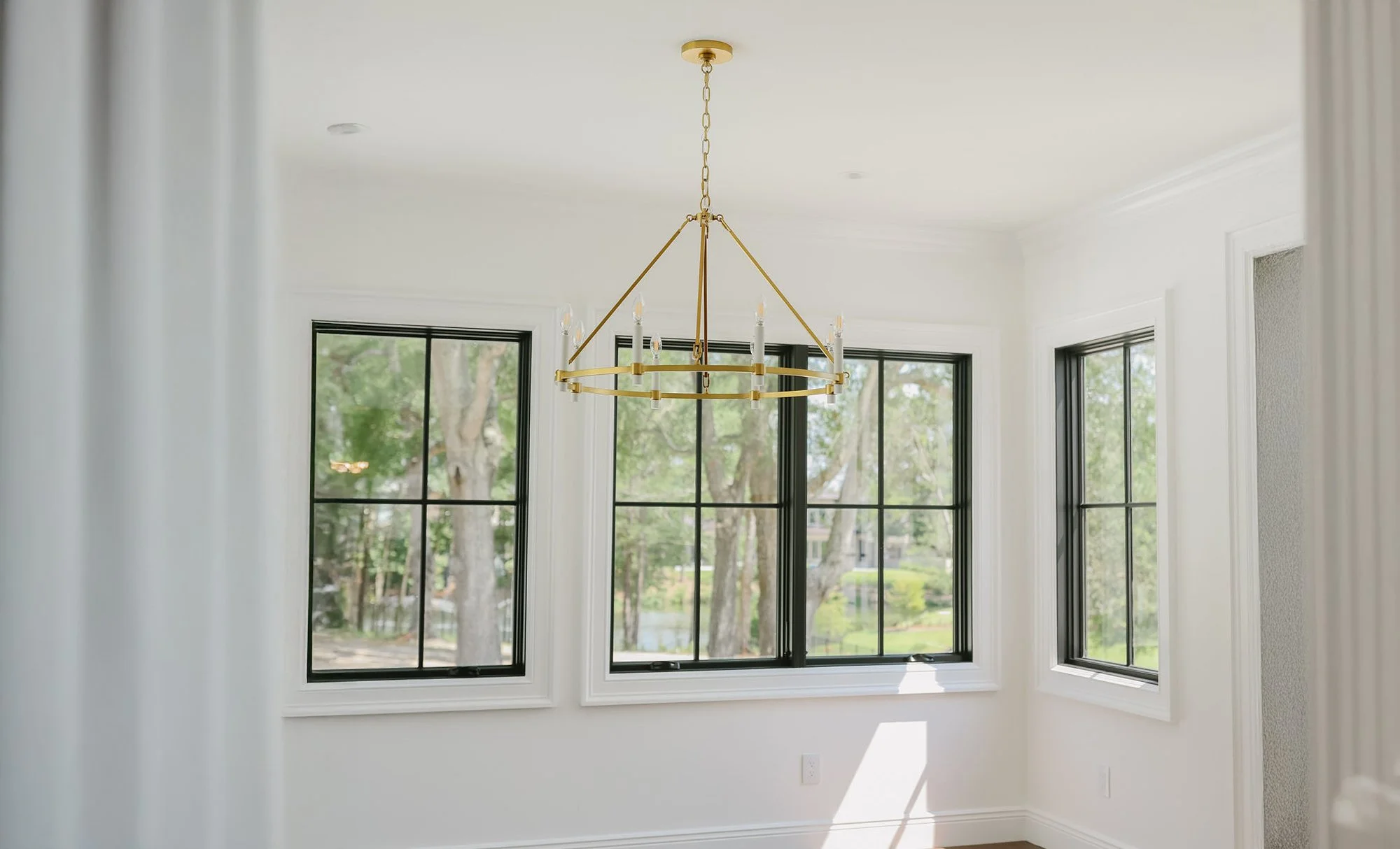 Bright room with large windows showing trees outside and a gold modern chandelier hanging from the ceiling.