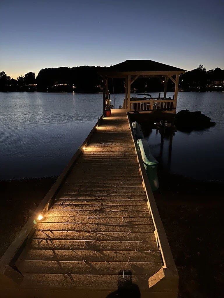 A wooden dock with built-in lights leading to a gazebo over the water at dusk, with trees and houses visible in the background.