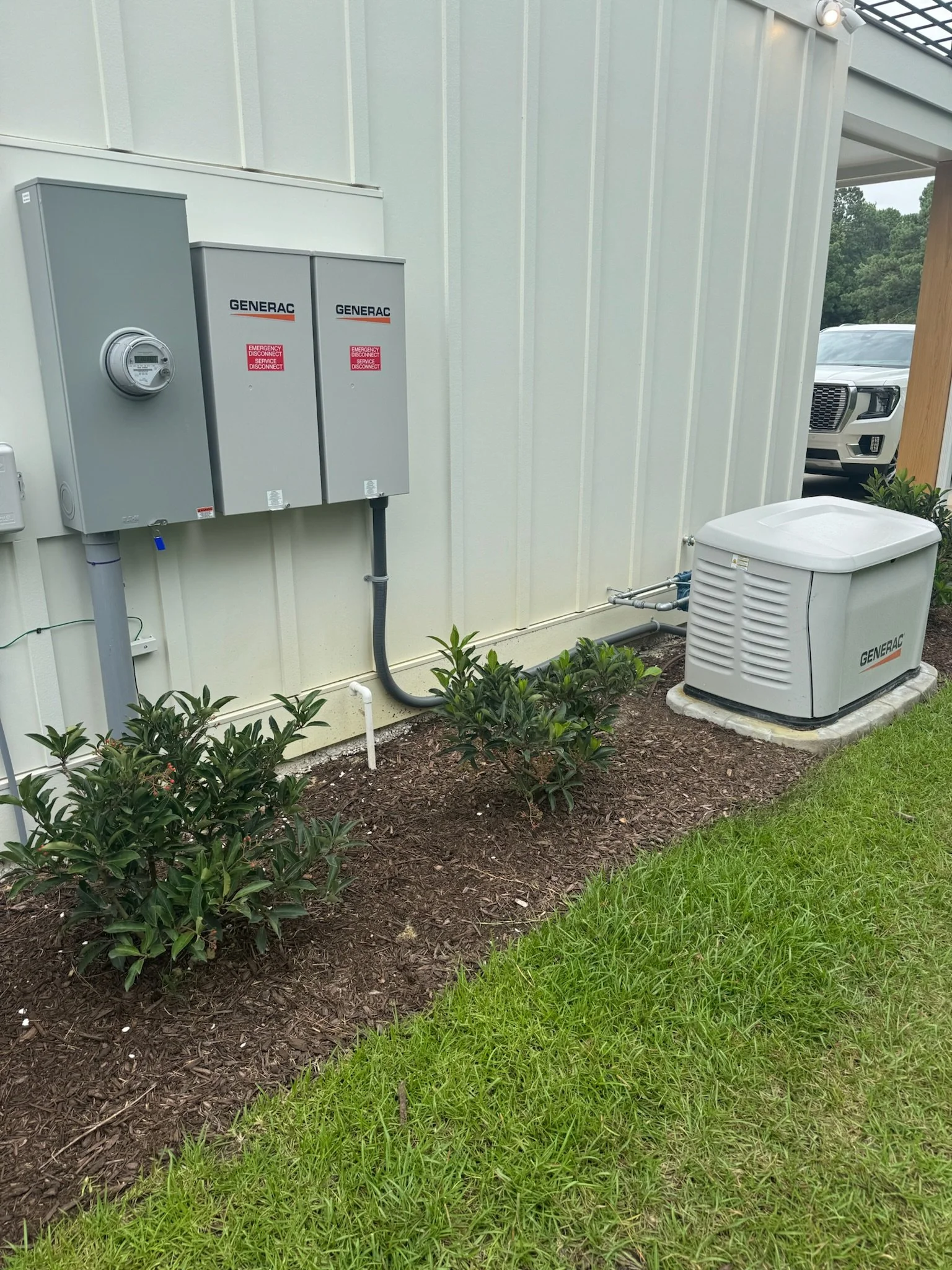 Exterior view of a building with two grey Generac generator cabinets, an electrical meter, and a white generator unit outside, with green grass and some bushes in the foreground.