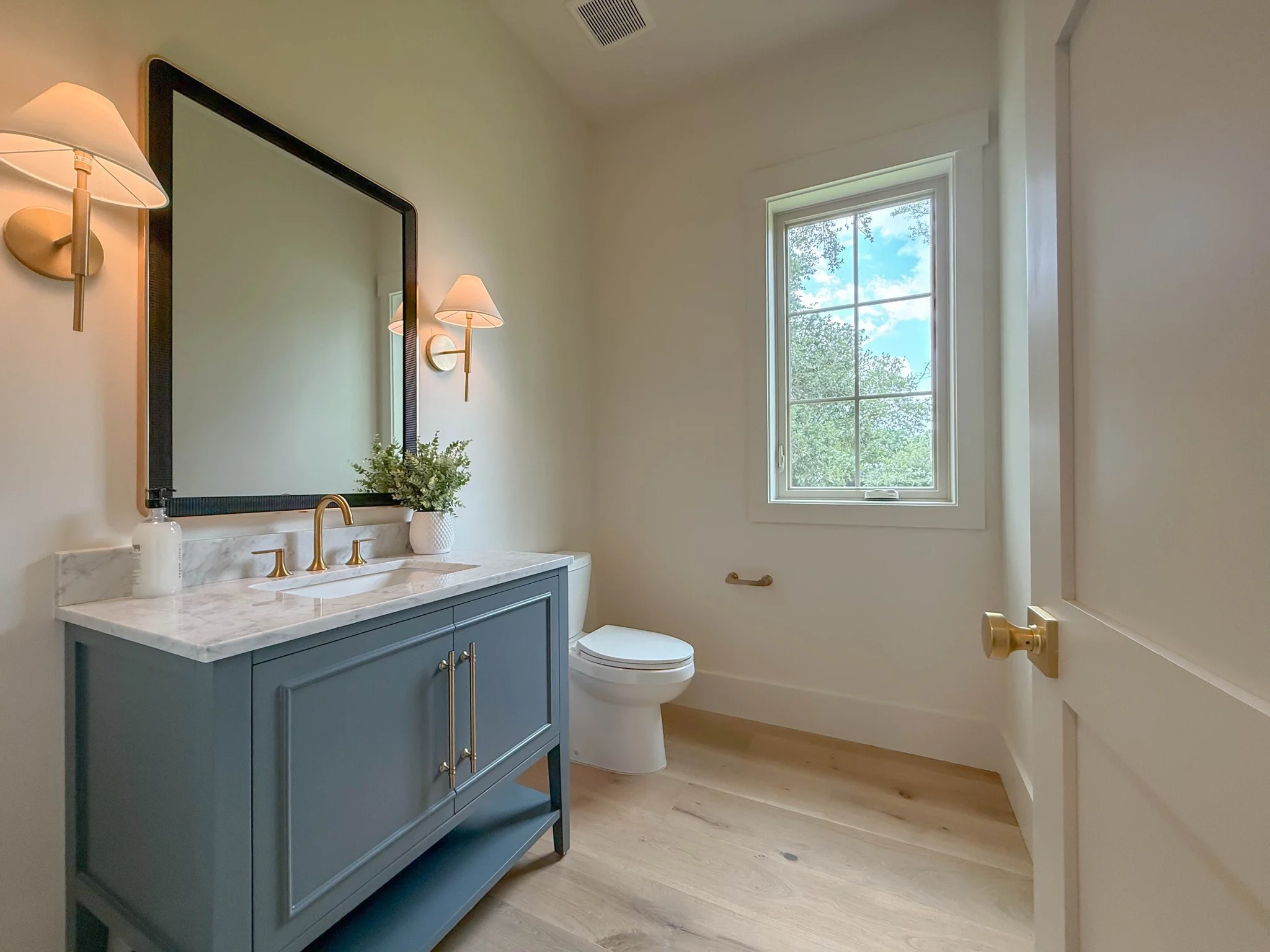 A modern bathroom featuring a blue vanity with a white marble countertop, a mirror, wall-mounted light fixtures, a potted plant, a window showing trees and a blue sky, and a toilet.