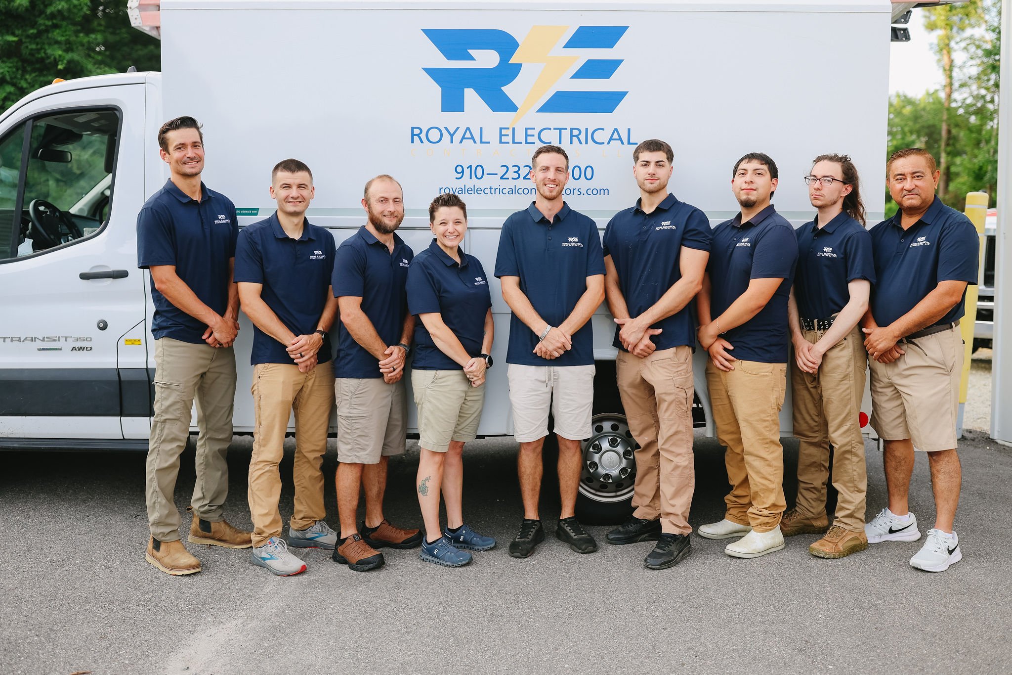 Group of nine people standing in front of a white Royal Electrical Contracting truck, outdoors, smiling at the camera. The group wears matching navy blue shirts with company logos, khaki pants, and various footwear. The truck features the company logo with a lightning bolt design and contact information.