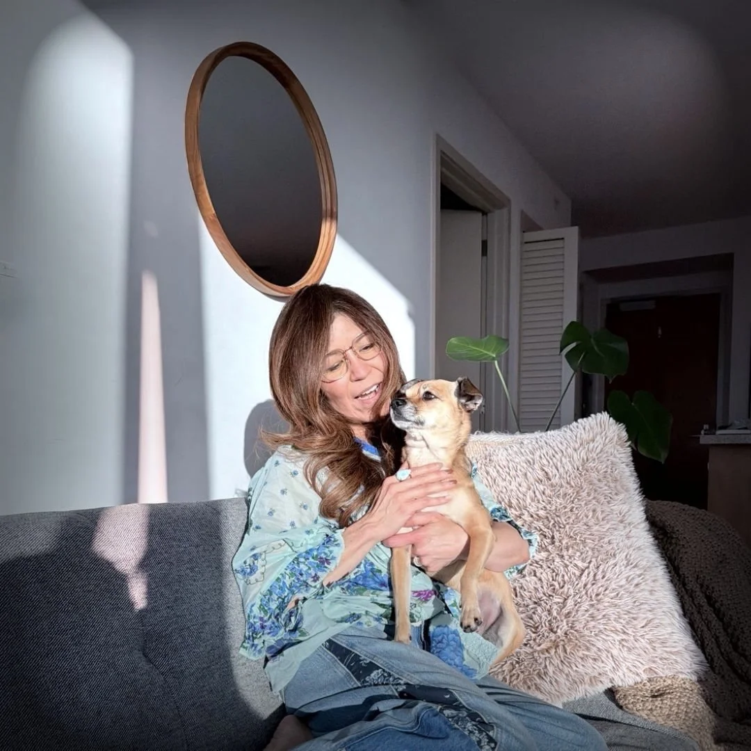 A woman with glasses sitting on a gray couch holding a small tan dog with black ears, in a living room with sunlight streaming through a window, a round wall mirror, and green houseplants.