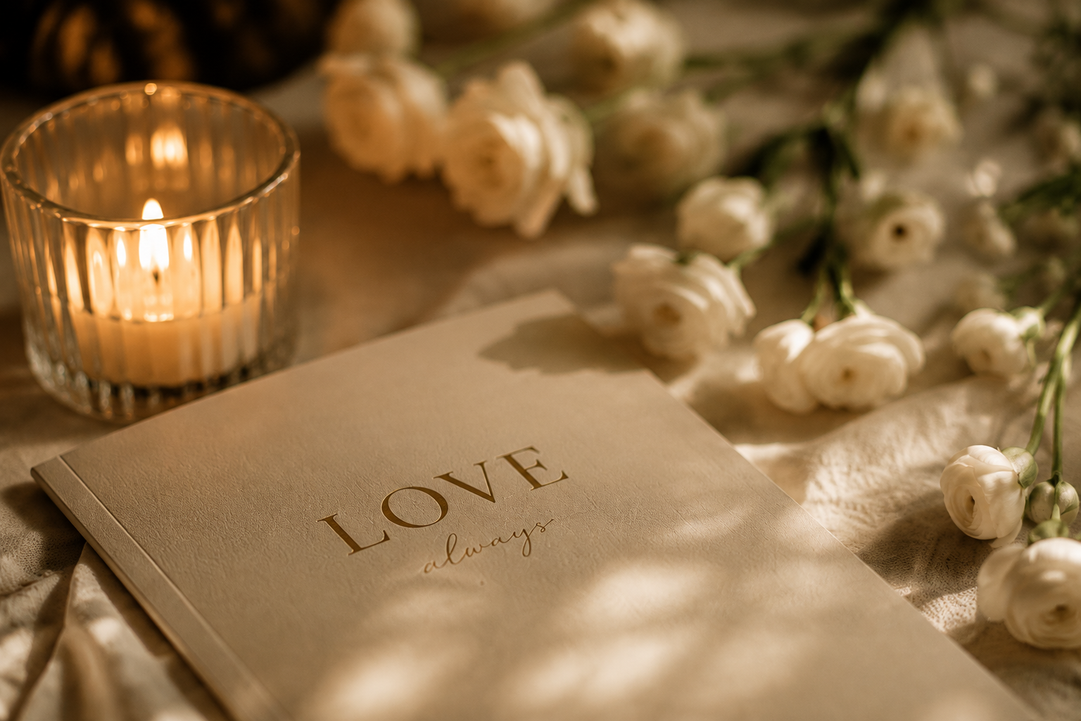 Kaart met de woorden 'LOVE always', witte bloemen en een brandende kaars op een tafel.