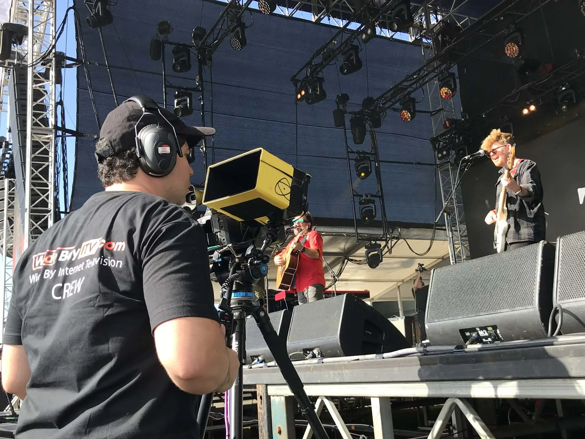 View of a stage setup with lighting rigs, a performer with blonde hair wearing glasses, and a guitarist in a red shirt playing in the background, with a camera operator in the foreground filming the performance.