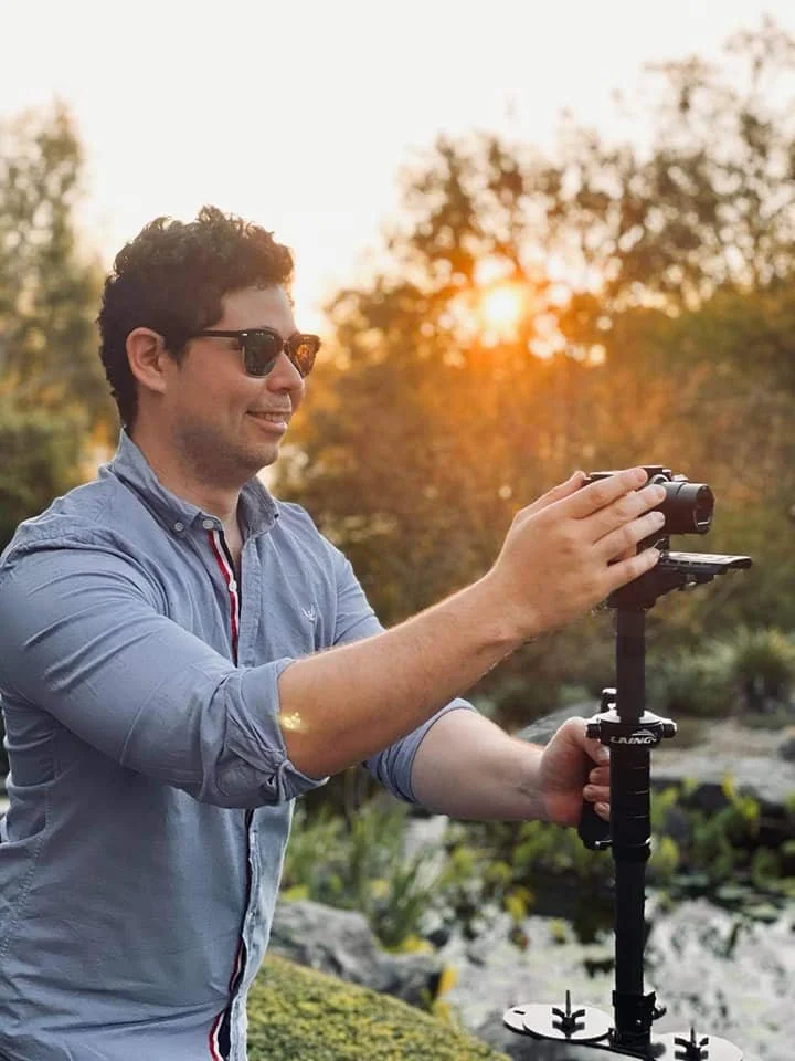 Man with sunglasses smiling outdoors at sunset, holding a camera on a stabilizer.