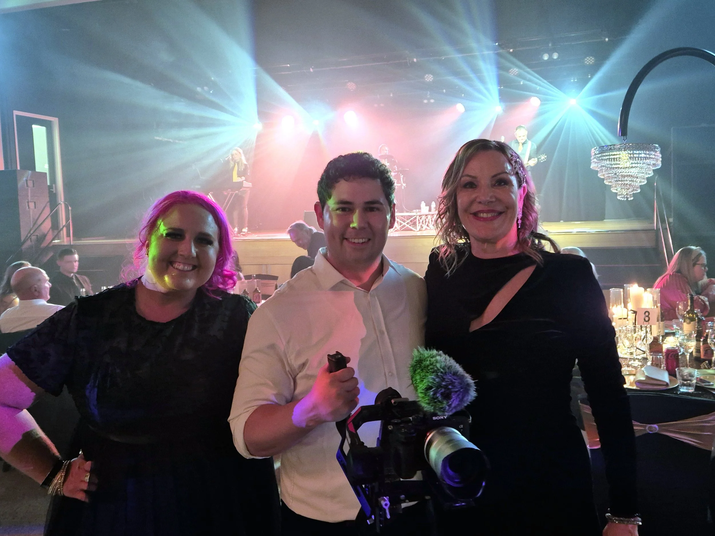 Three people smiling and posing for a photo at a lively event with a band performing on stage in the background, vibrant lighting, and seated guests at tables.