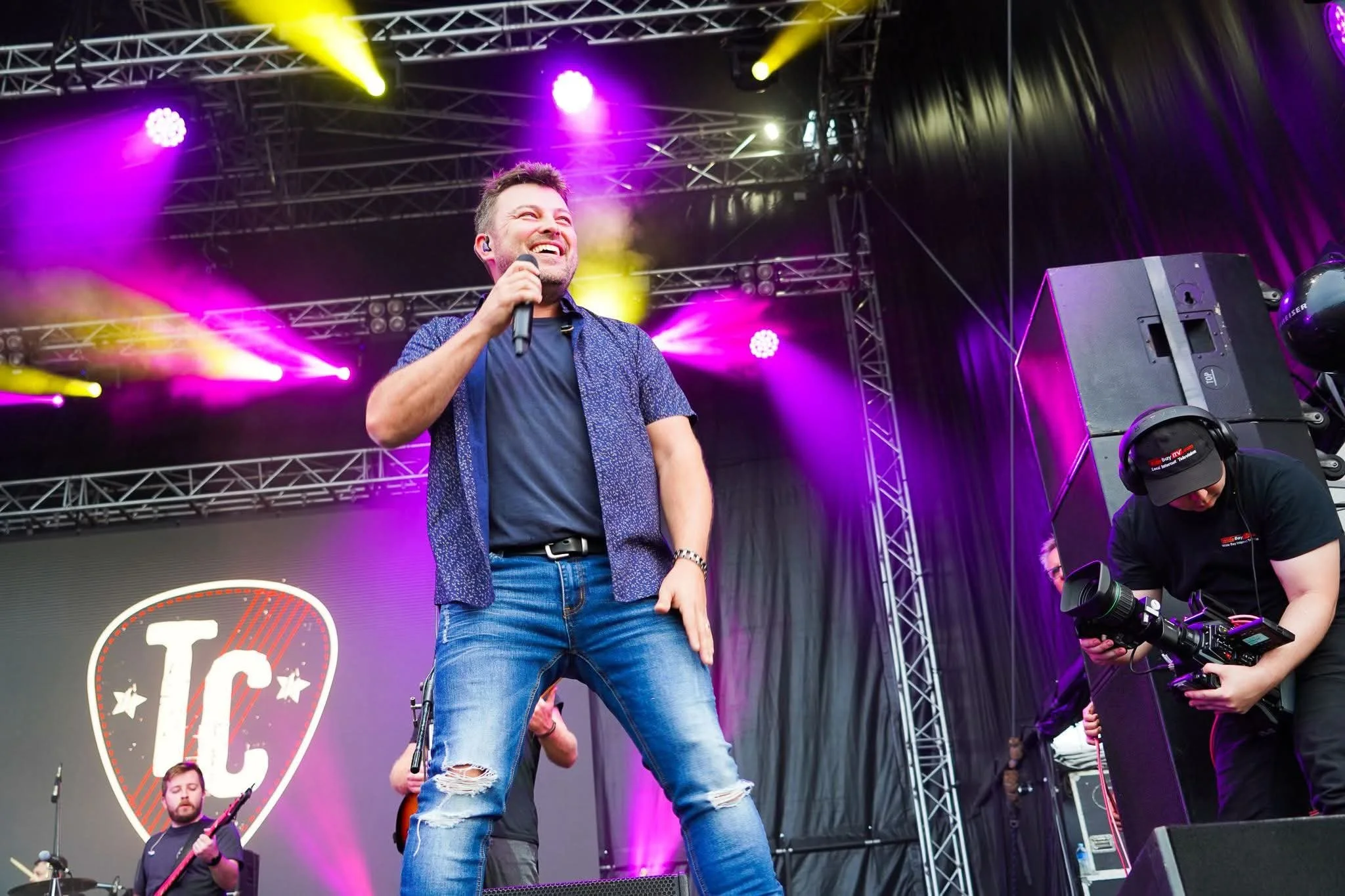 A man holding a microphone on stage at a concert or music event, smiling. Behind him are colorful stage lights and other band members, with a large speaker and a sign with the letter 'T' visible.